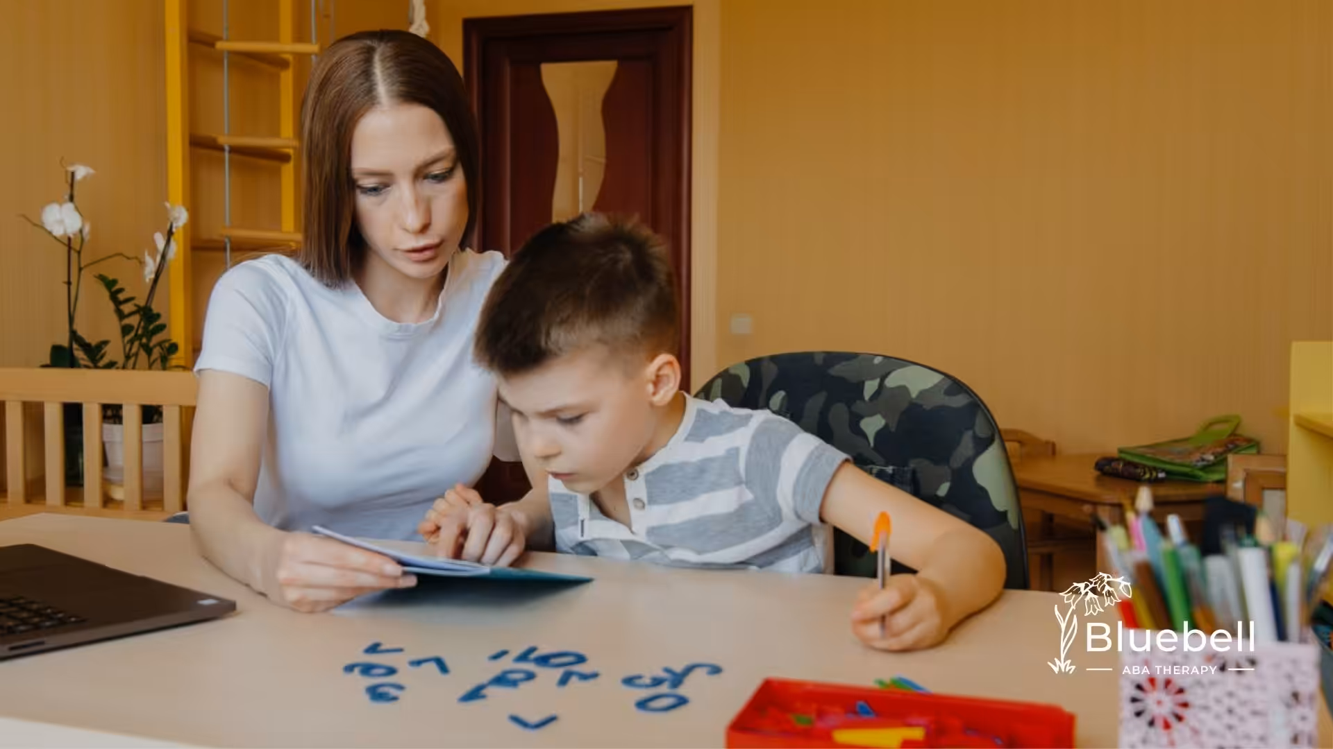 ABA therapist guiding young boy in a learning activity at a desk with educational tools.