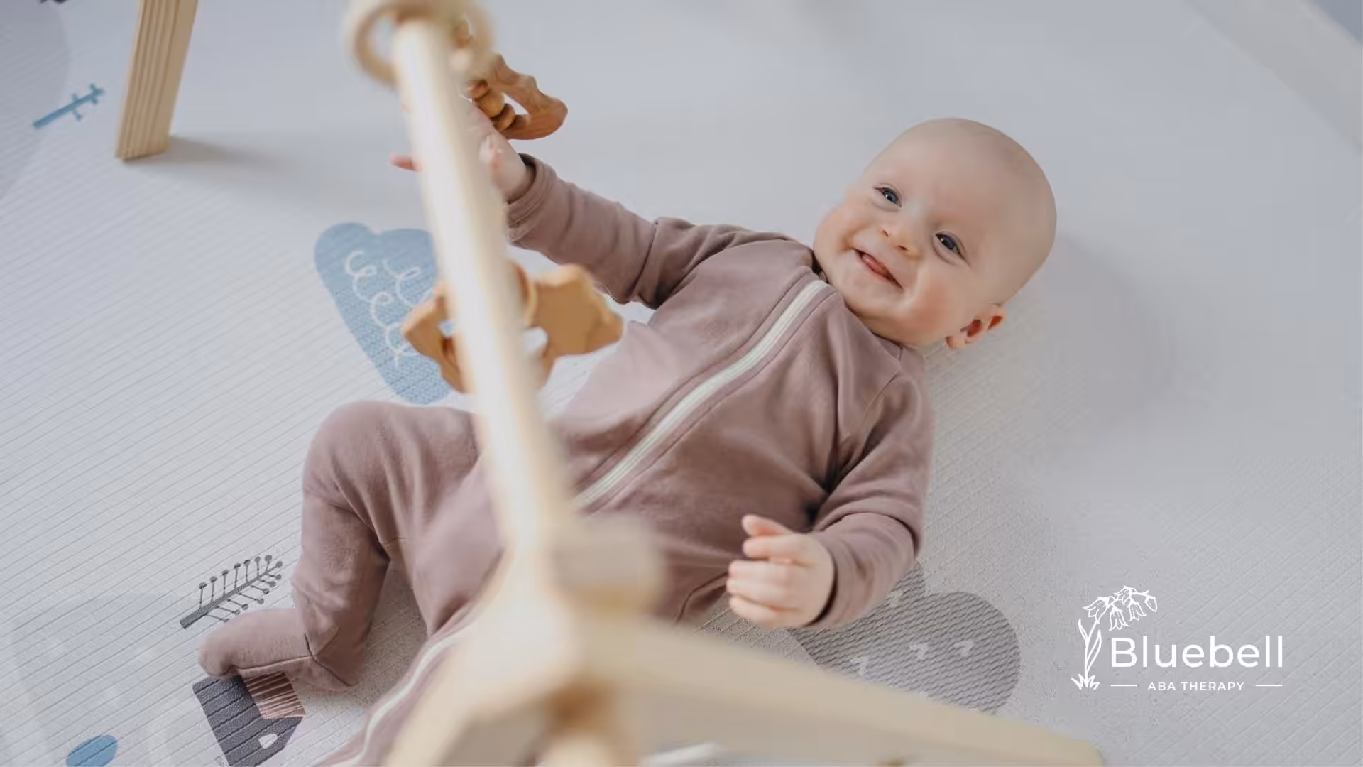 Smiling autistic baby in a soft brown outfit playing on a white mat with a wooden baby gym in NC.