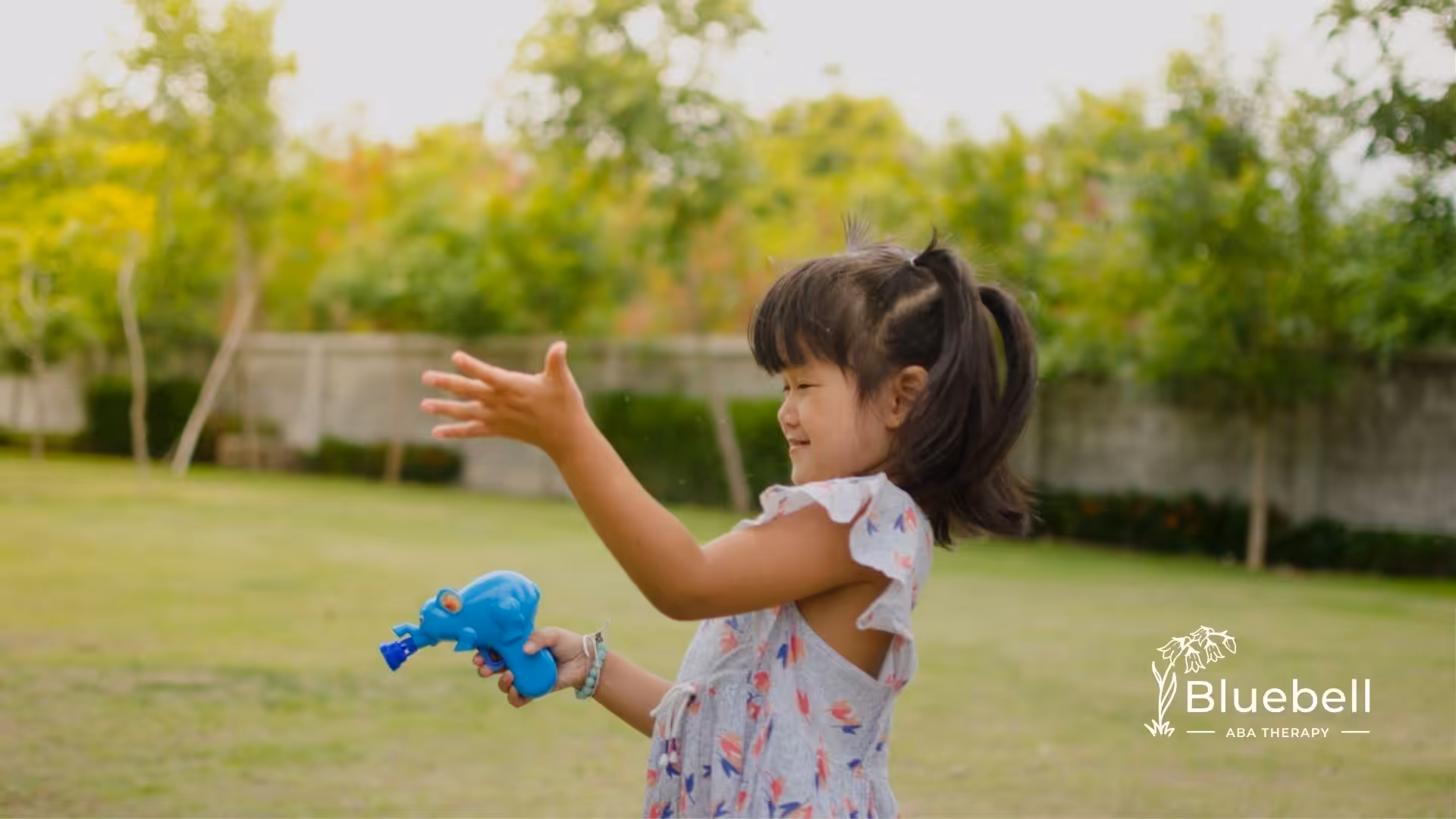 A smiling girl playing outdoors with a blue water gun in a sunny backyard.