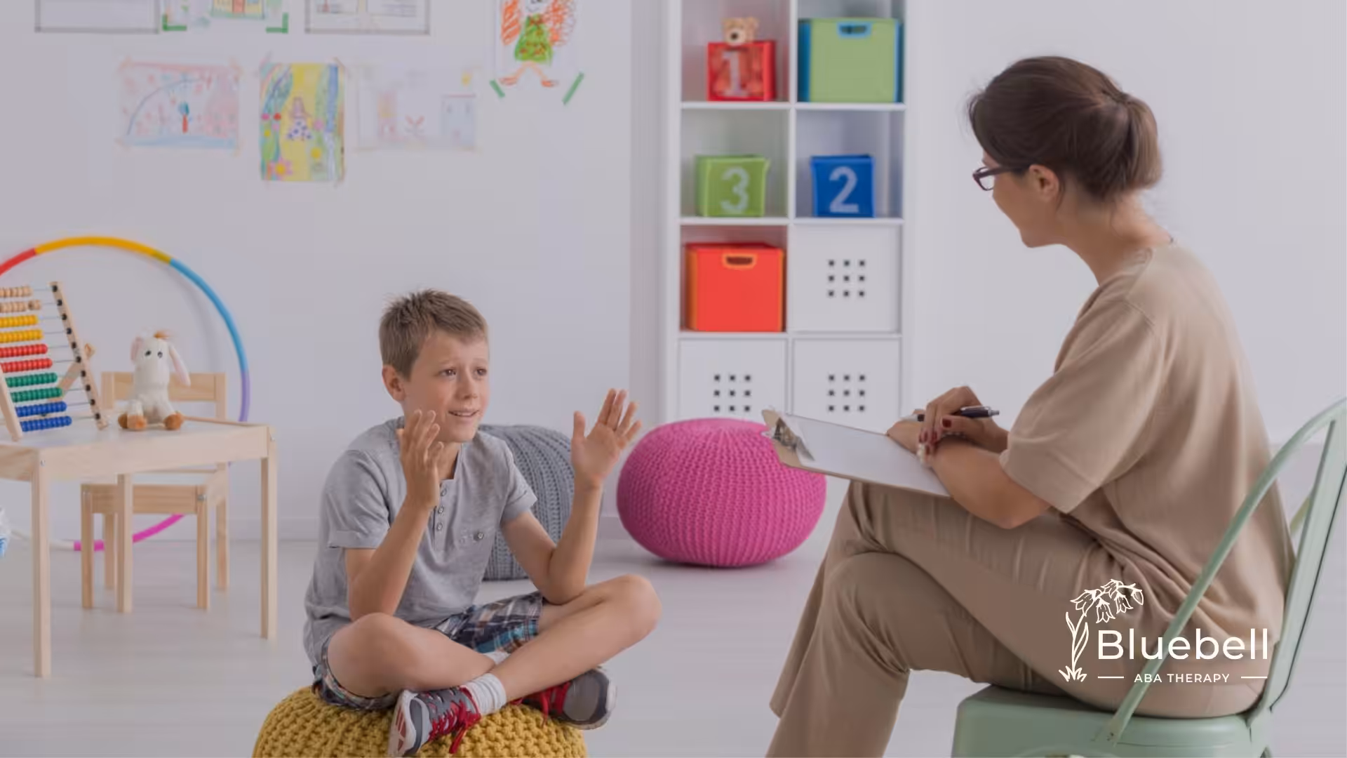 A boy sitting on a pouf in a bright classroom, talking to an ABA therapist taking notes.