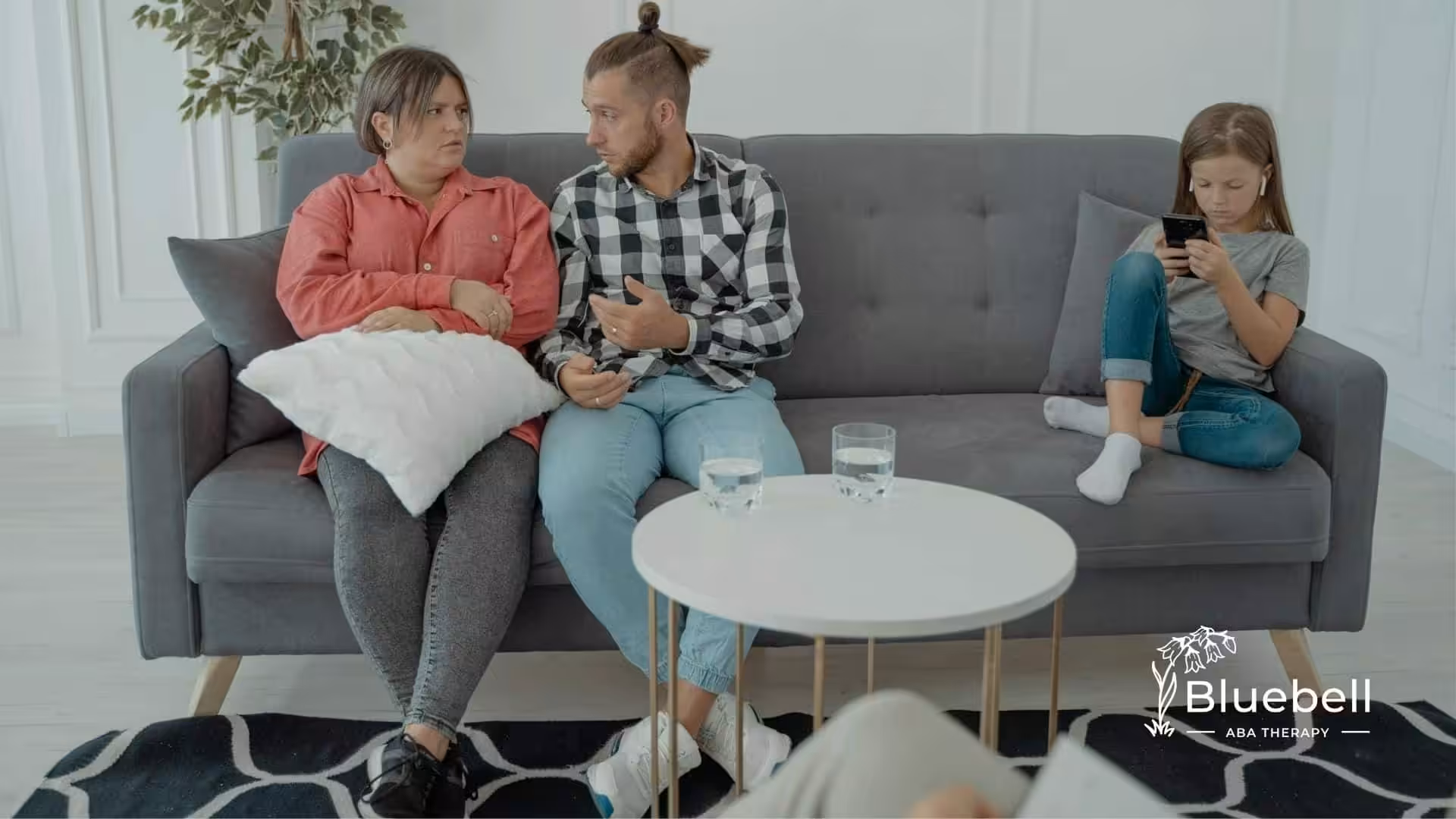 Parents discussing what questions to ask to ABA therapist while autistic daughter plays on a phone.