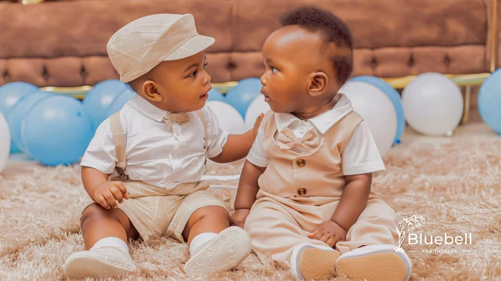 Two autistic babies dressed in beige formal outfits, sitting on a rug, looking at each other in NC.