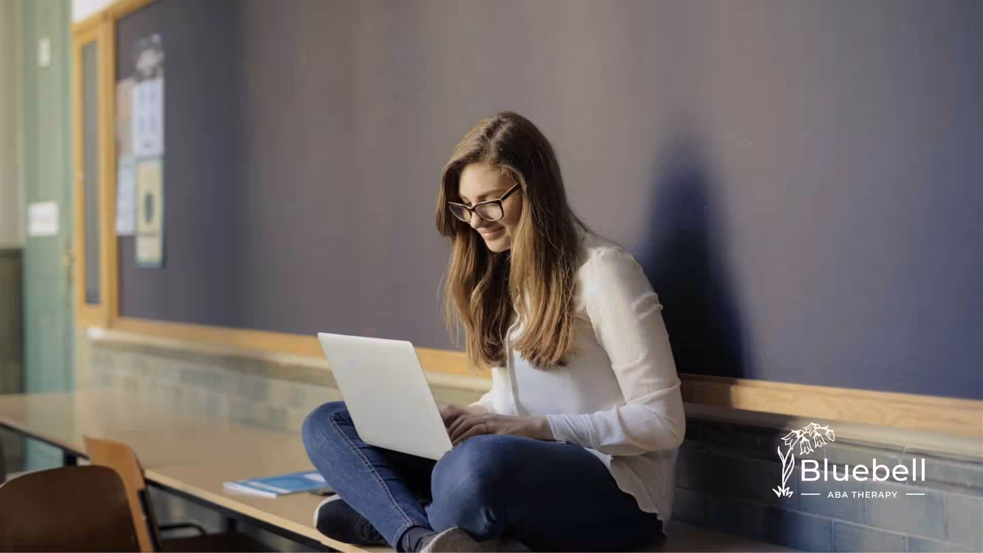 A BCBA sitting on a bench in a classroom, working on her laptop with a smile in North Carolina.
