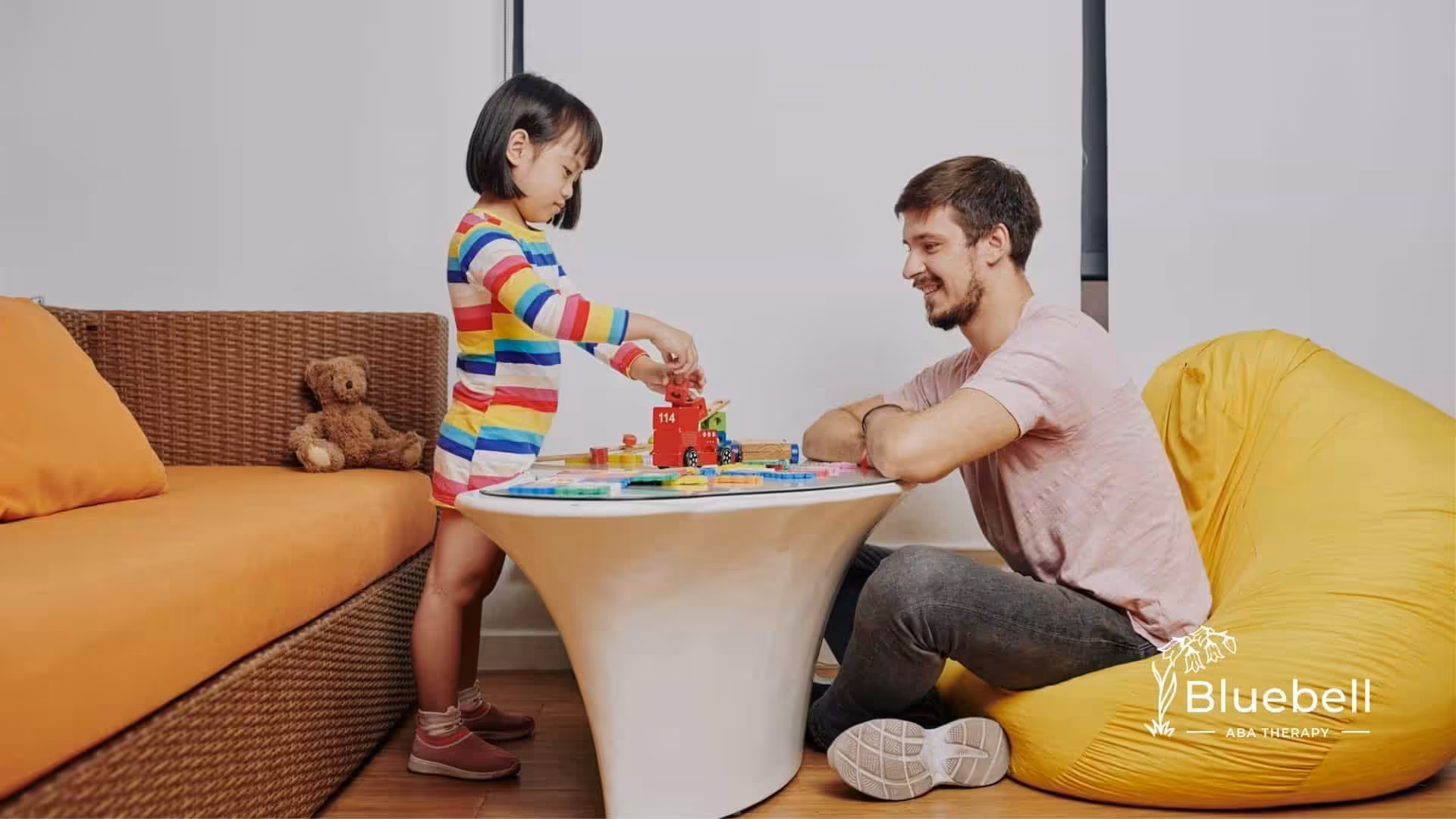 A littlw builds with toy blocks while an ABA therapist observes from a beanbag chair.