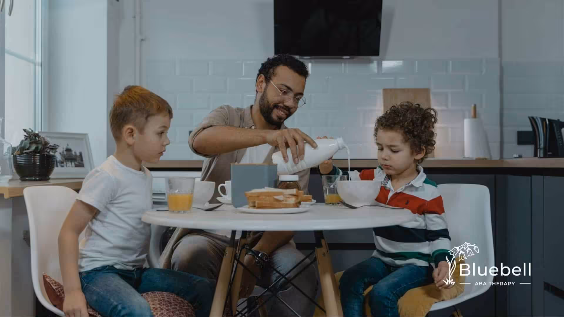 Father pouring milk into autistic son’s cereal bowl at breakfast with another child watching in NC.