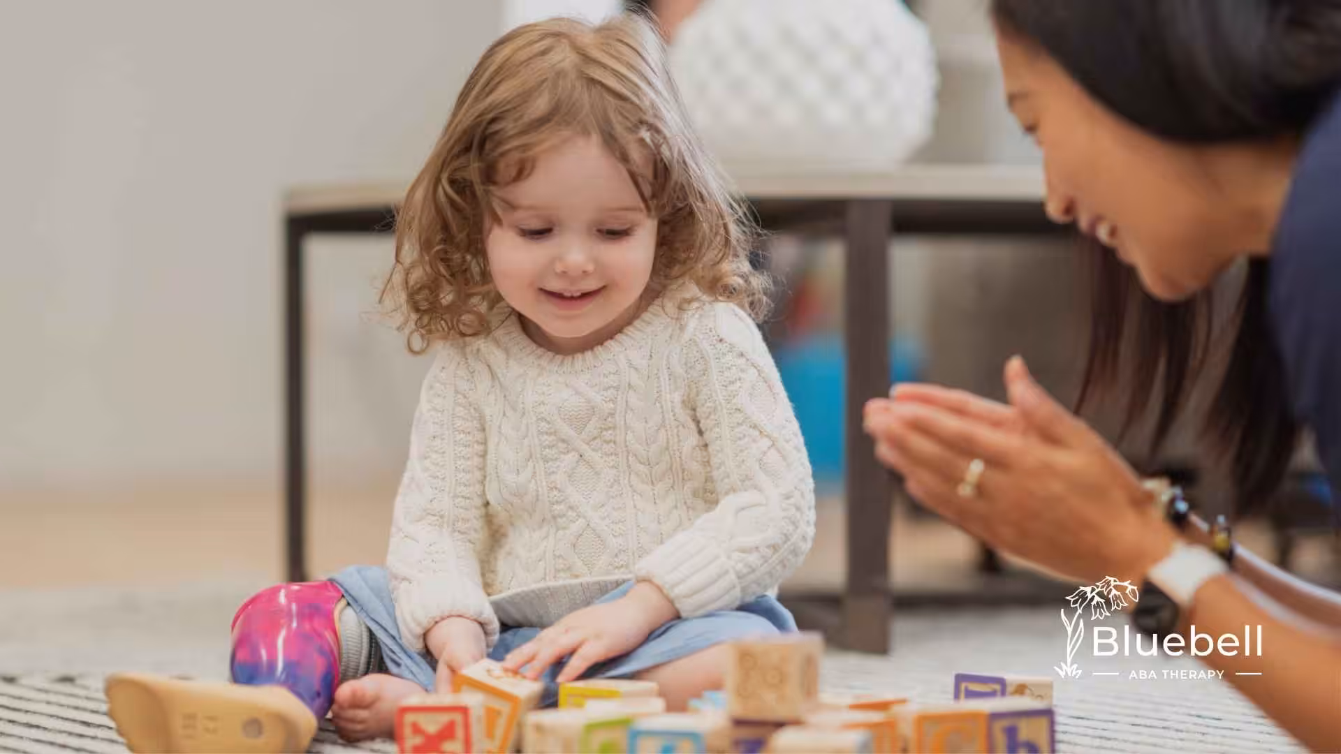 A smiling child with curly hair and a colorful prosthetic leg plays with blocks on a rug, while an adult claps encouragingly nearby. Warm, supportive atmosphere.