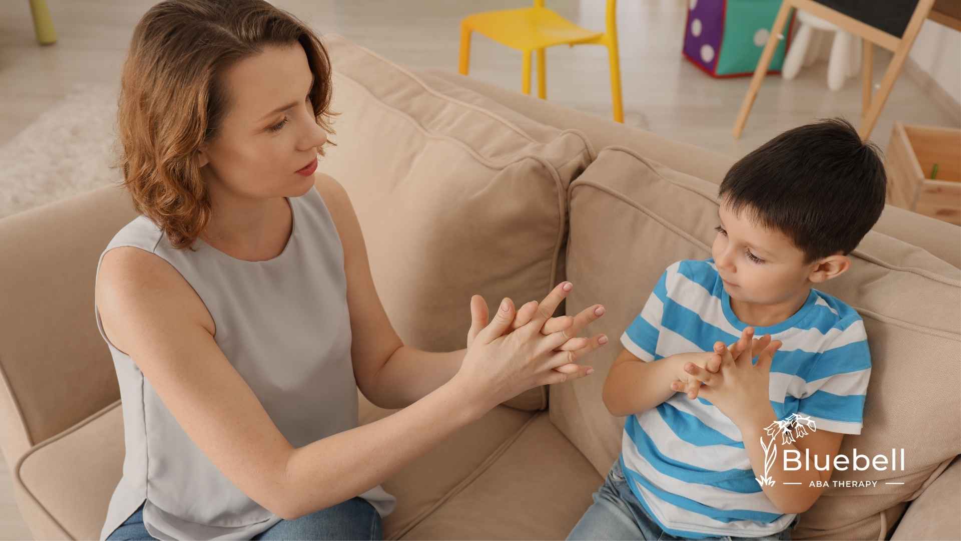 A woman and a young boy sit on a beige couch, engaged in a focused hand exercise. The setting is bright and calm.