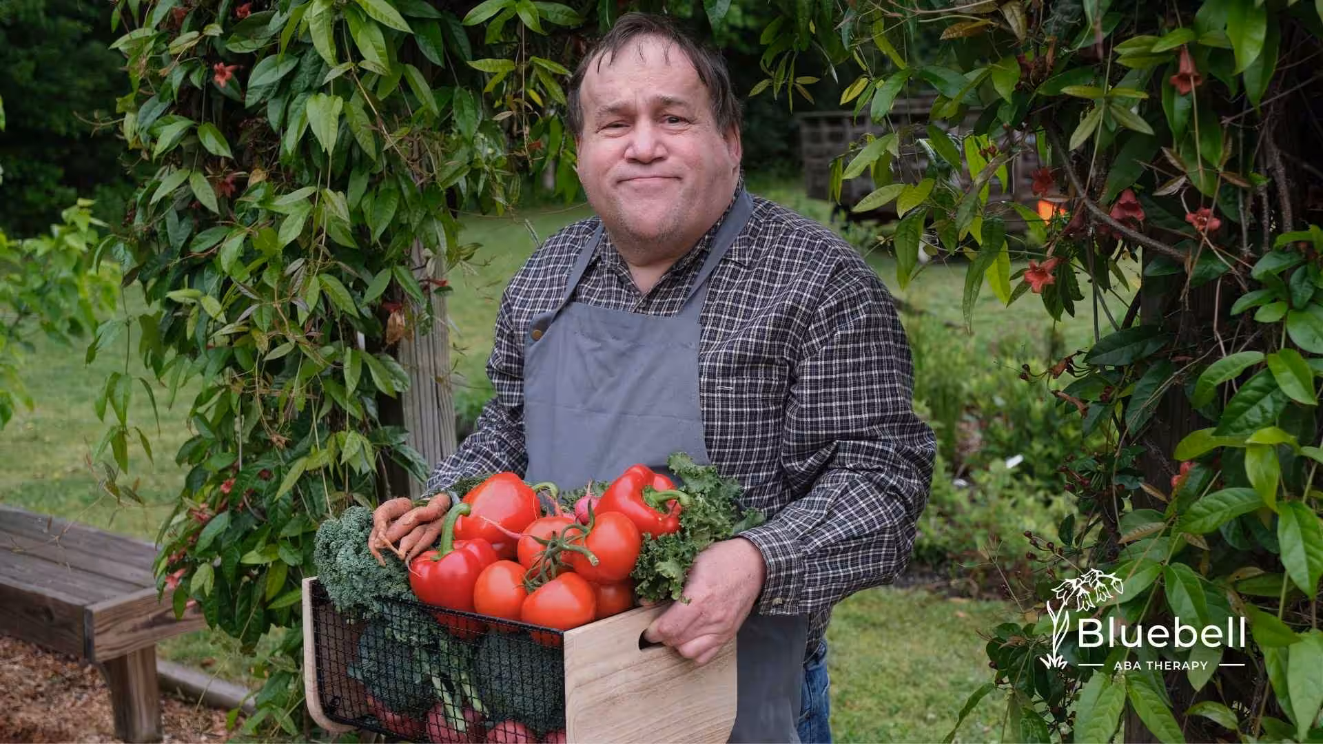 Man in a garden under a leafy archway, wearing a plaid shirt and apron, holds a crate of fresh vegetables, conveying a sense of pride and contentment.
