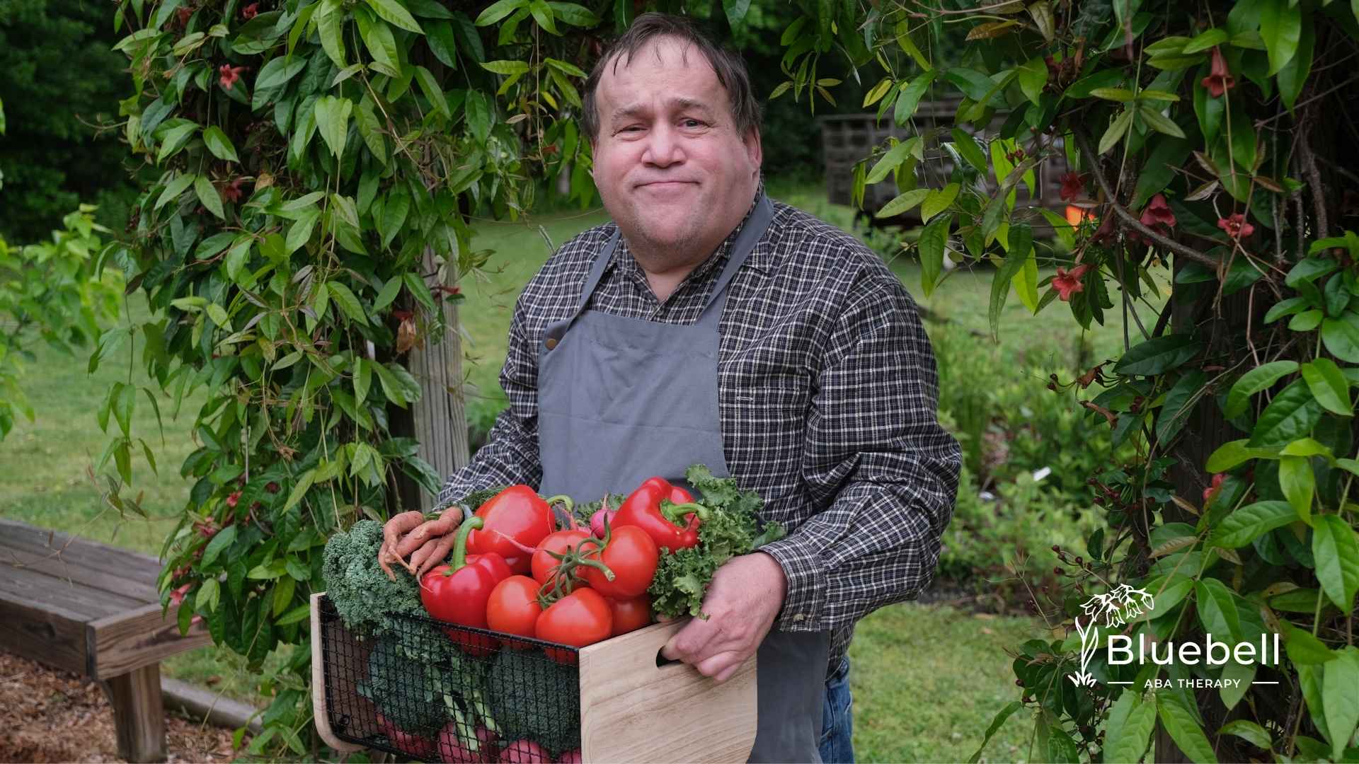 Man in a garden under a leafy archway, wearing a plaid shirt and apron, holds a crate of fresh vegetables, conveying a sense of pride and contentment.