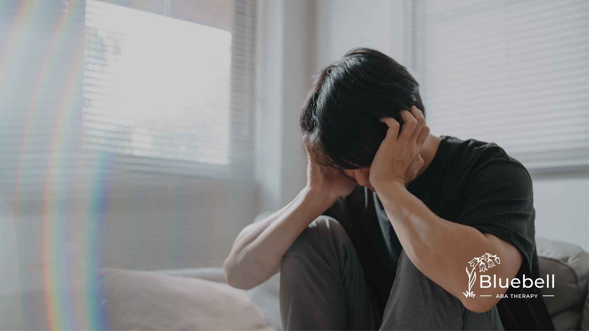 A person sits on a bed in a sunlit room, holding their head in distress. A subtle rainbow reflection appears. The image conveys a sense of anxiety or overwhelm.