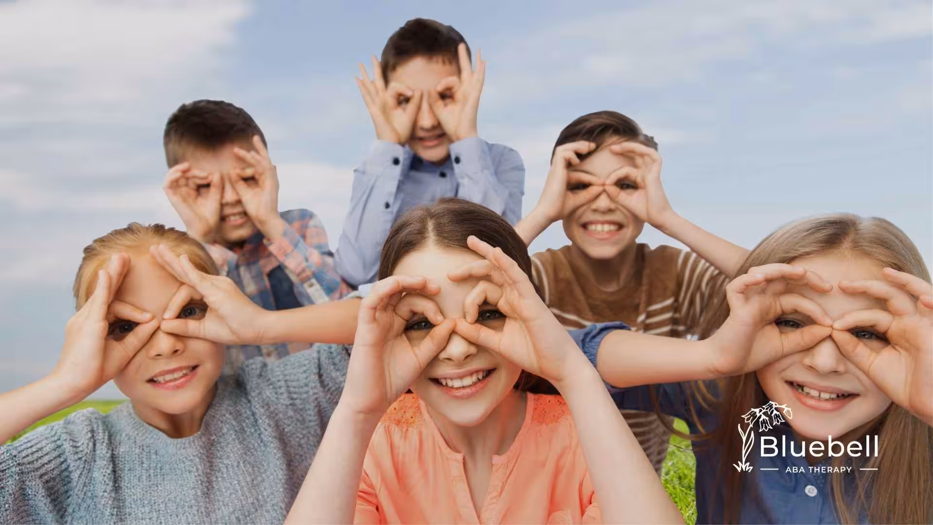 Six children smiling and making hand circles over their eyes outdoors under a blue sky. The mood is playful and joyful.