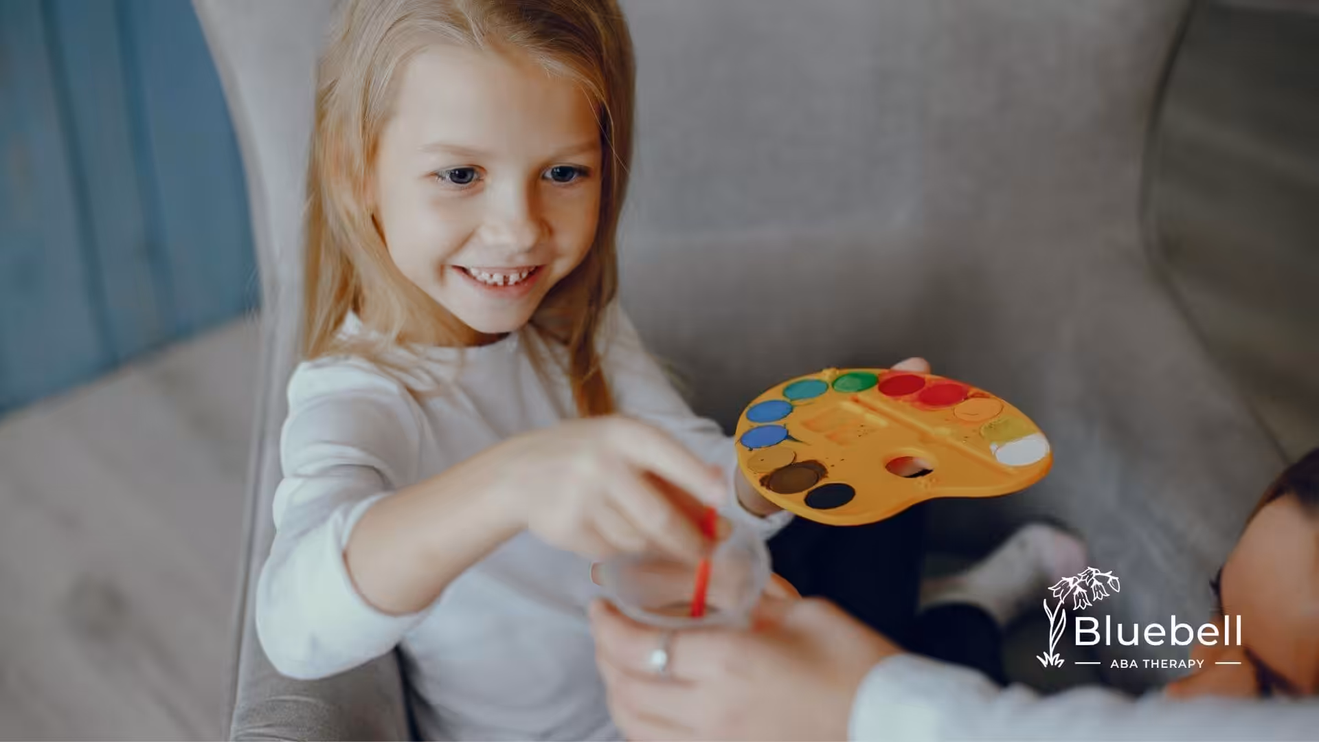A girl with autism smiling while holding a paint palette and dipping a brush into water.