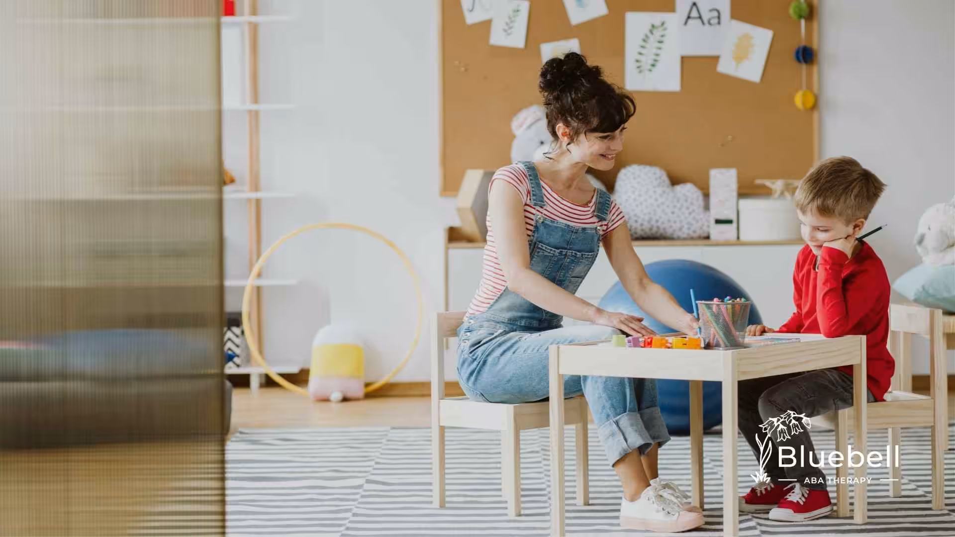 A woman in overalls smiles while sitting at a table with a young boy in a red shirt. They are engaged in a playful activity in a colorful classroom.