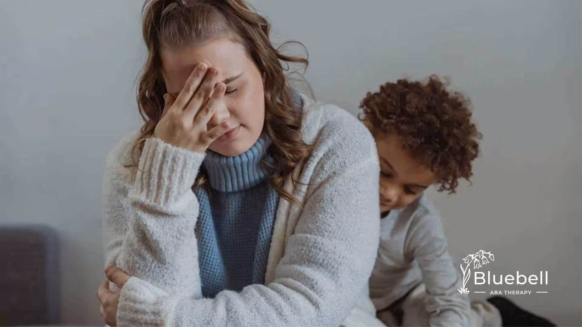 A stressed woman holds her head while a child with autism hugs her from behind.