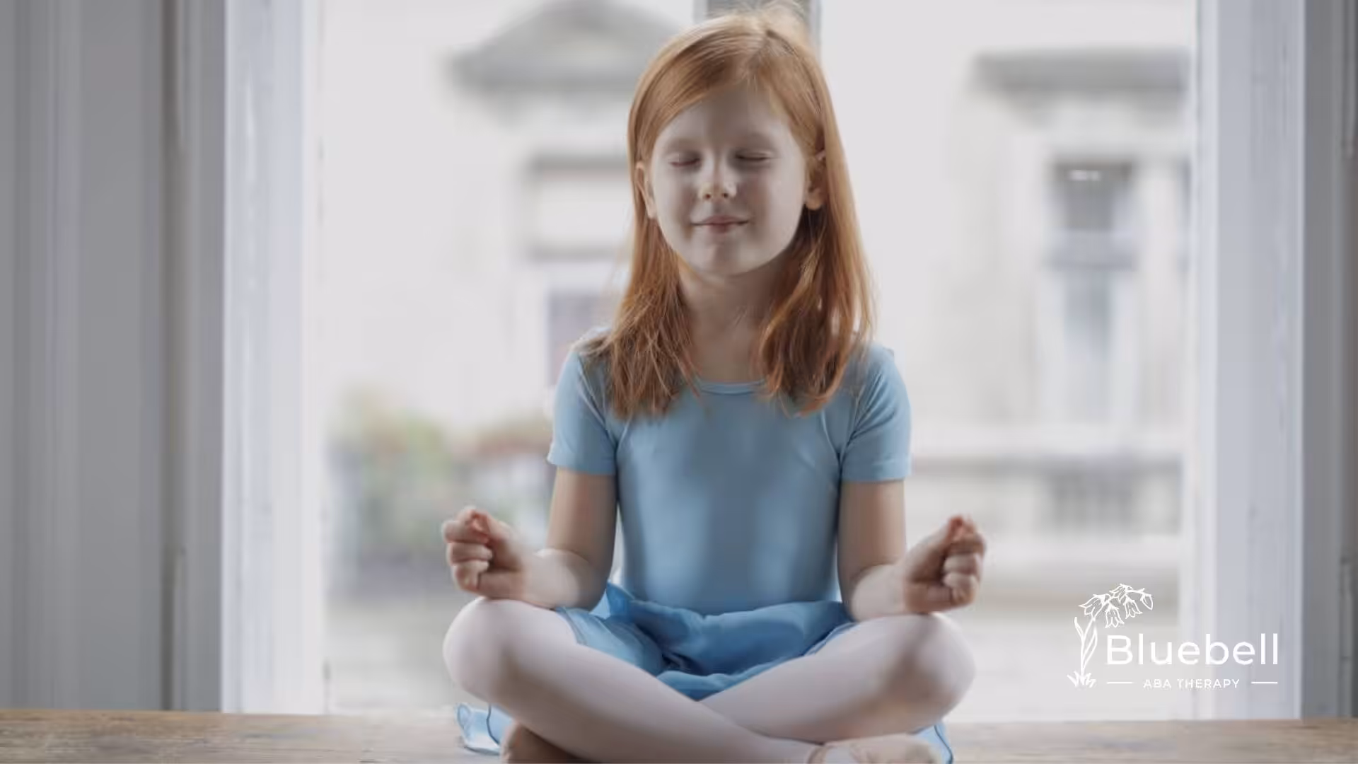 A red-haired girl in a blue dress meditates cross-legged by a window during ABA therapy.