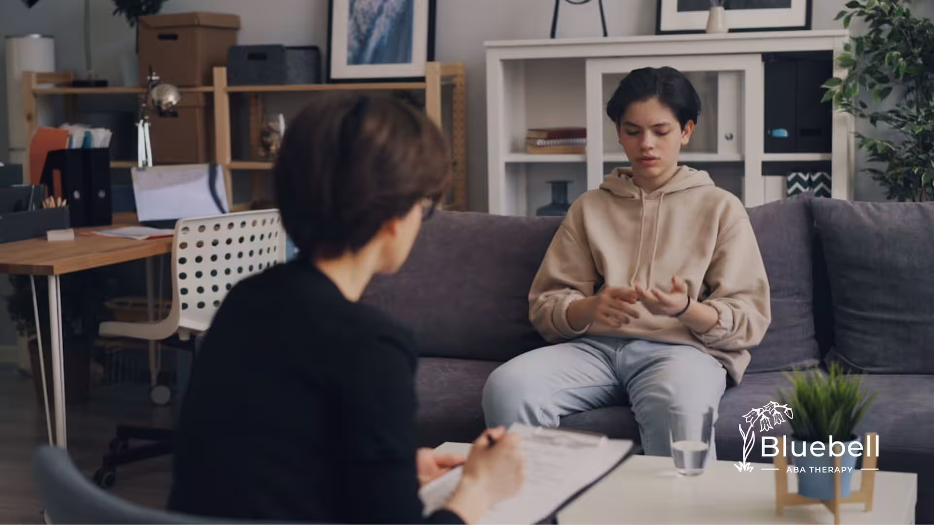 A teenage boy with autism sits on a couch talking to an ABA therapist holding a clipboard.