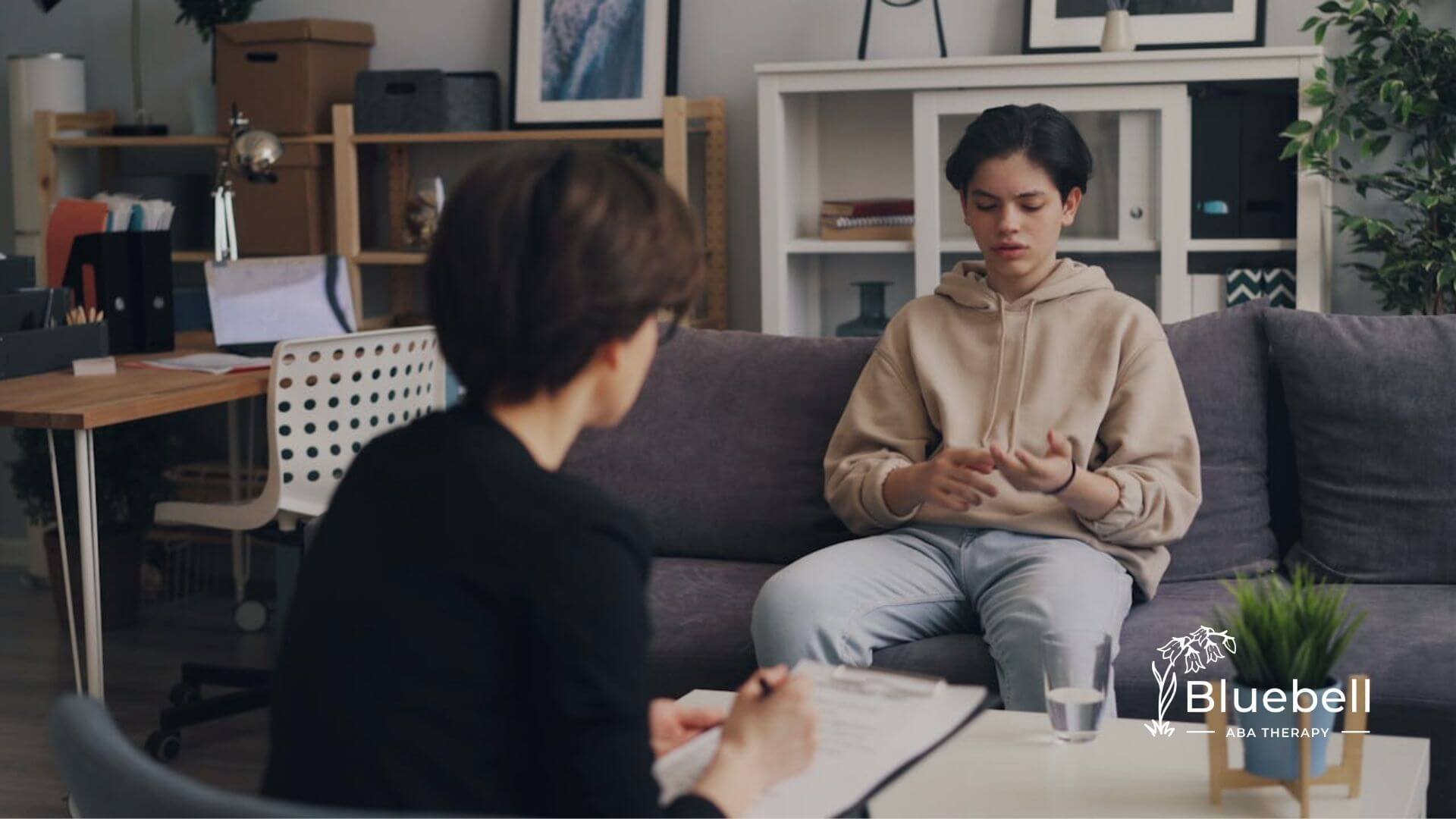 A teenage boy with autism sits on a couch talking to an ABA therapist holding a clipboard.