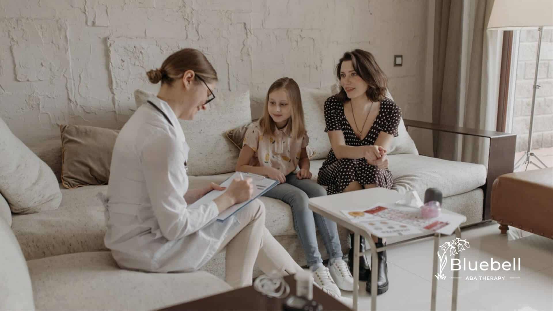An ABA therapist takes notes while talking with a girl and her mother on a couch in ABA therapy.