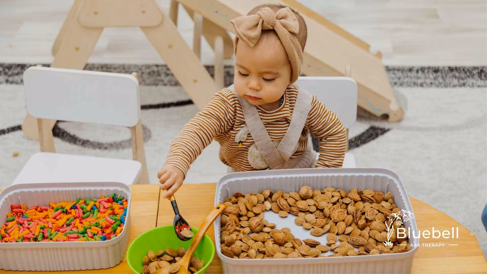 A toddler in a striped outfit, wearing a bow headband, plays with sensory bins. One bin has colorful pasta, the other has nuts. The setting is a wooden play area.