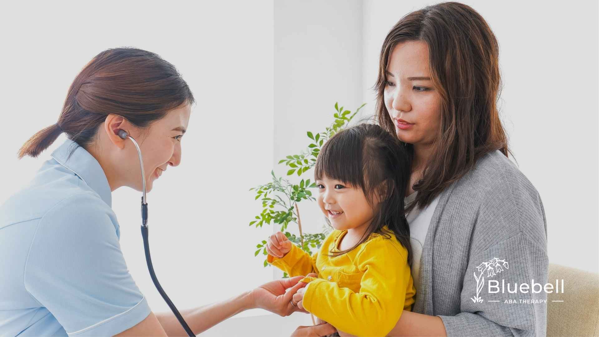 A smiling nurse with a stethoscope interacts with a young child and mother. The child, in a yellow shirt, appears happy. A plant is in the background.