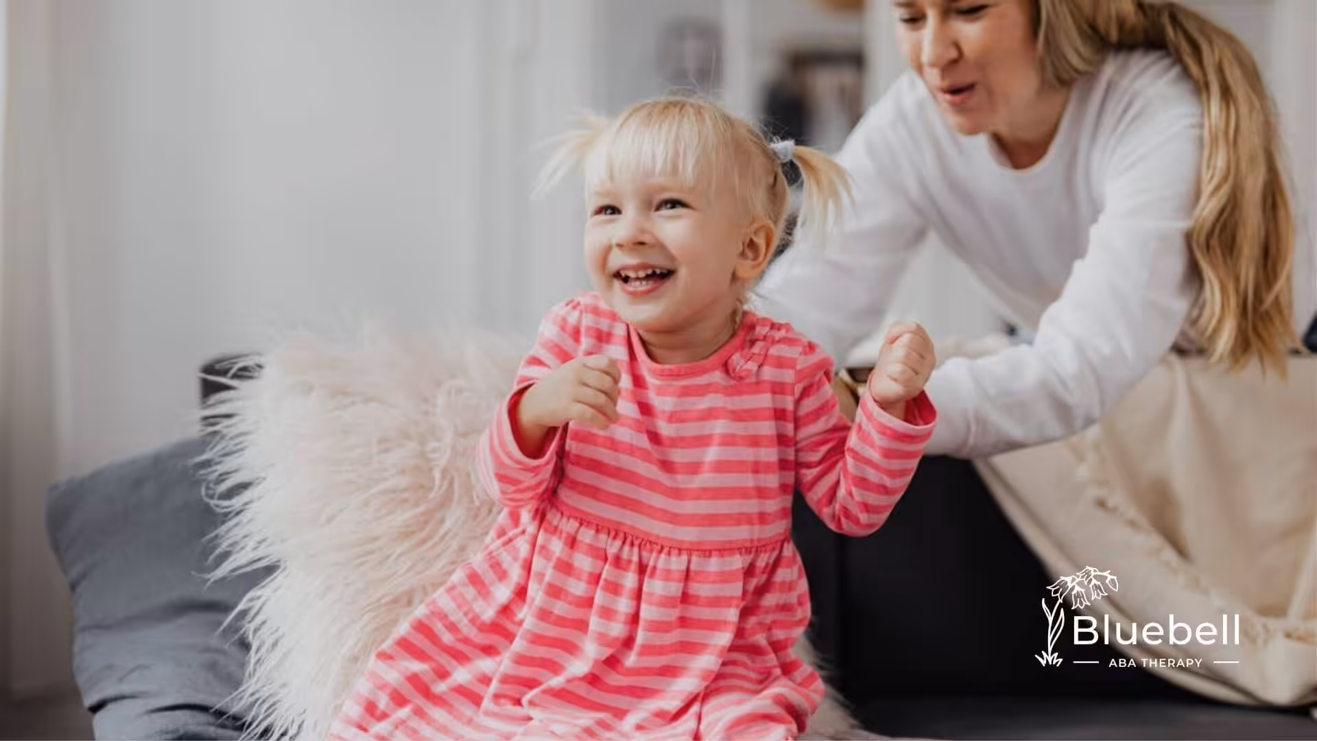 A laughing child in a pink striped dress sits on a couch with a BCBA in ABA therapy.
