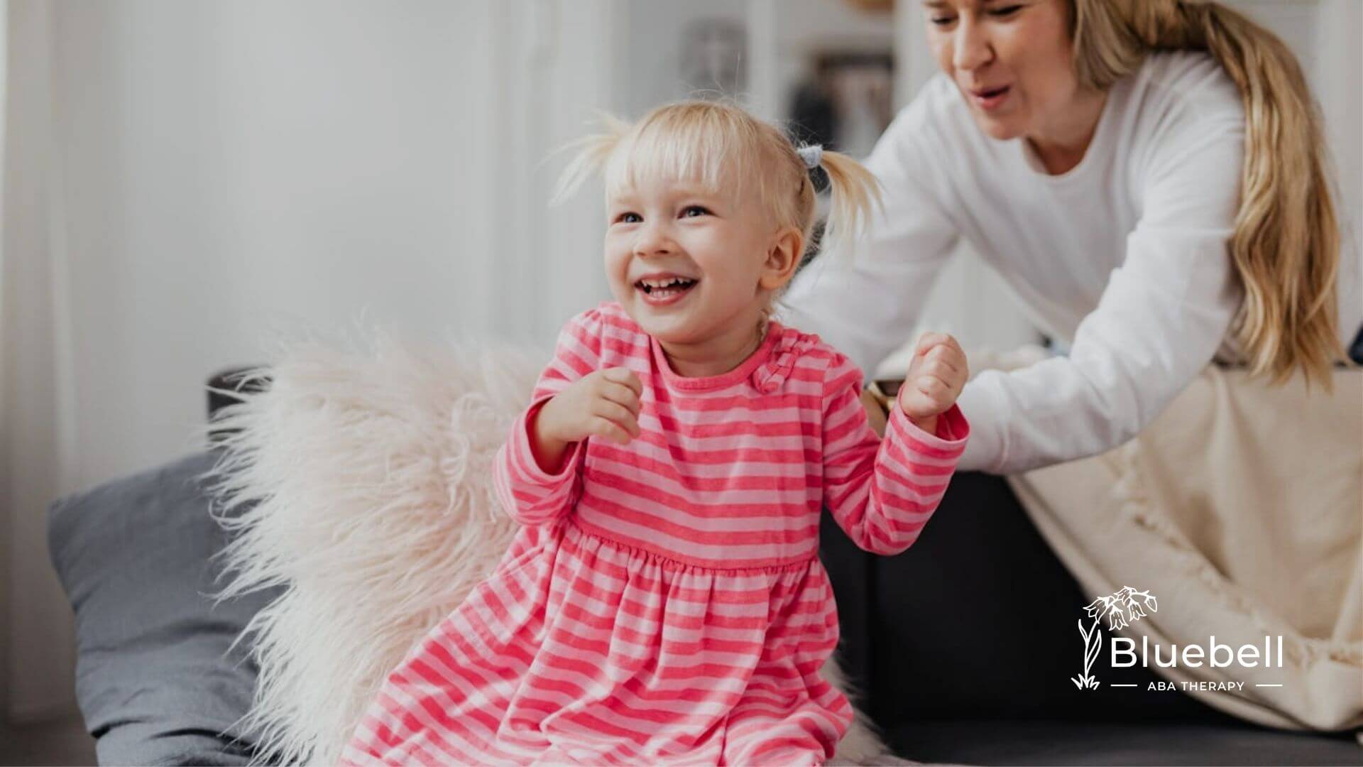 A laughing child in a pink striped dress sits on a couch with a BCBA in ABA therapy.