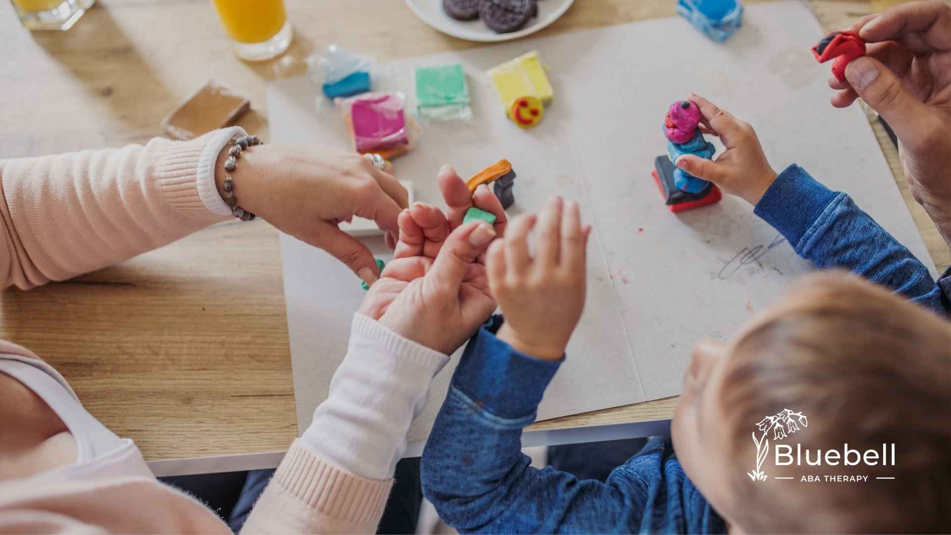 A child and two adults mold colorful clay on a table, conveying creativity and learning. The atmosphere is warm and engaging, with hands shown actively shaping the clay.