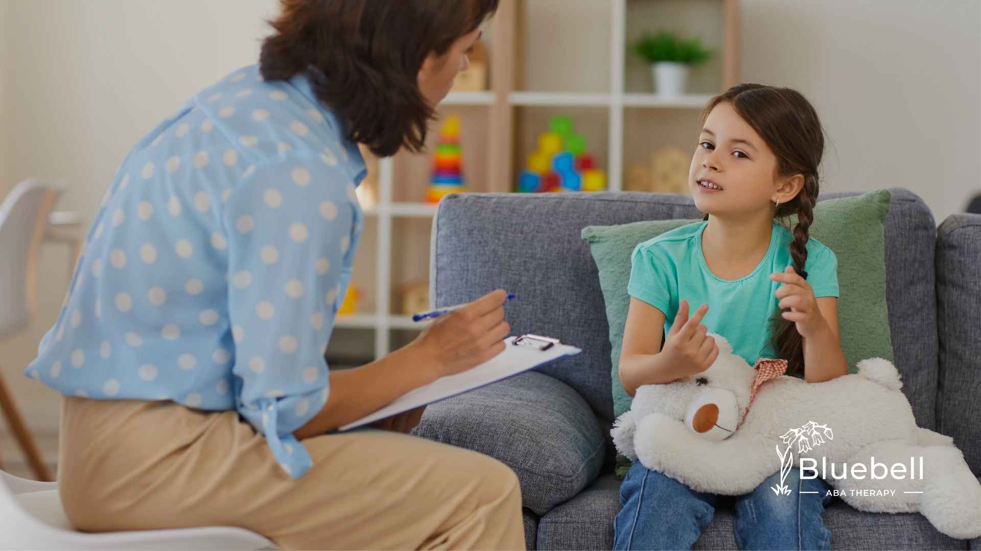 A therapist in a blue polka dot shirt takes notes while talking to a young girl holding a teddy bear. They sit on a gray sofa in a cozy, colorful playroom.