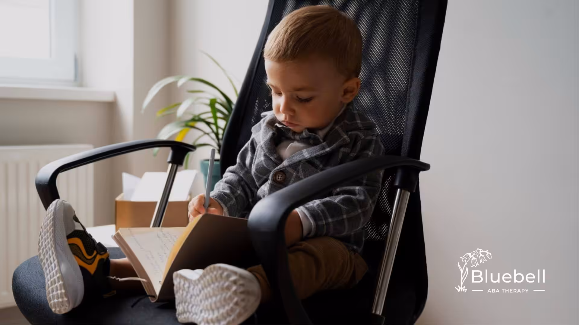 A toddler with autism dressed in a checked suit writes in a notebook while seated in an office chair.