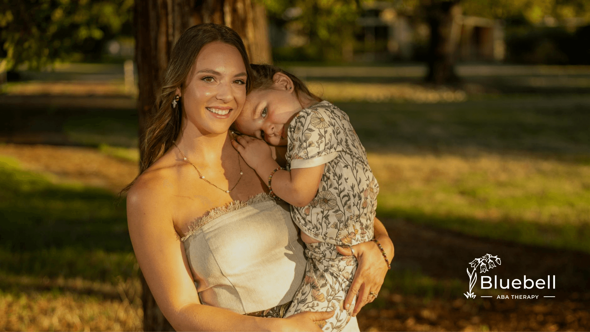 ABA therapist embraces the autistic child in sunny outdoor setting, showing warmth, closeness, and gentle affection.