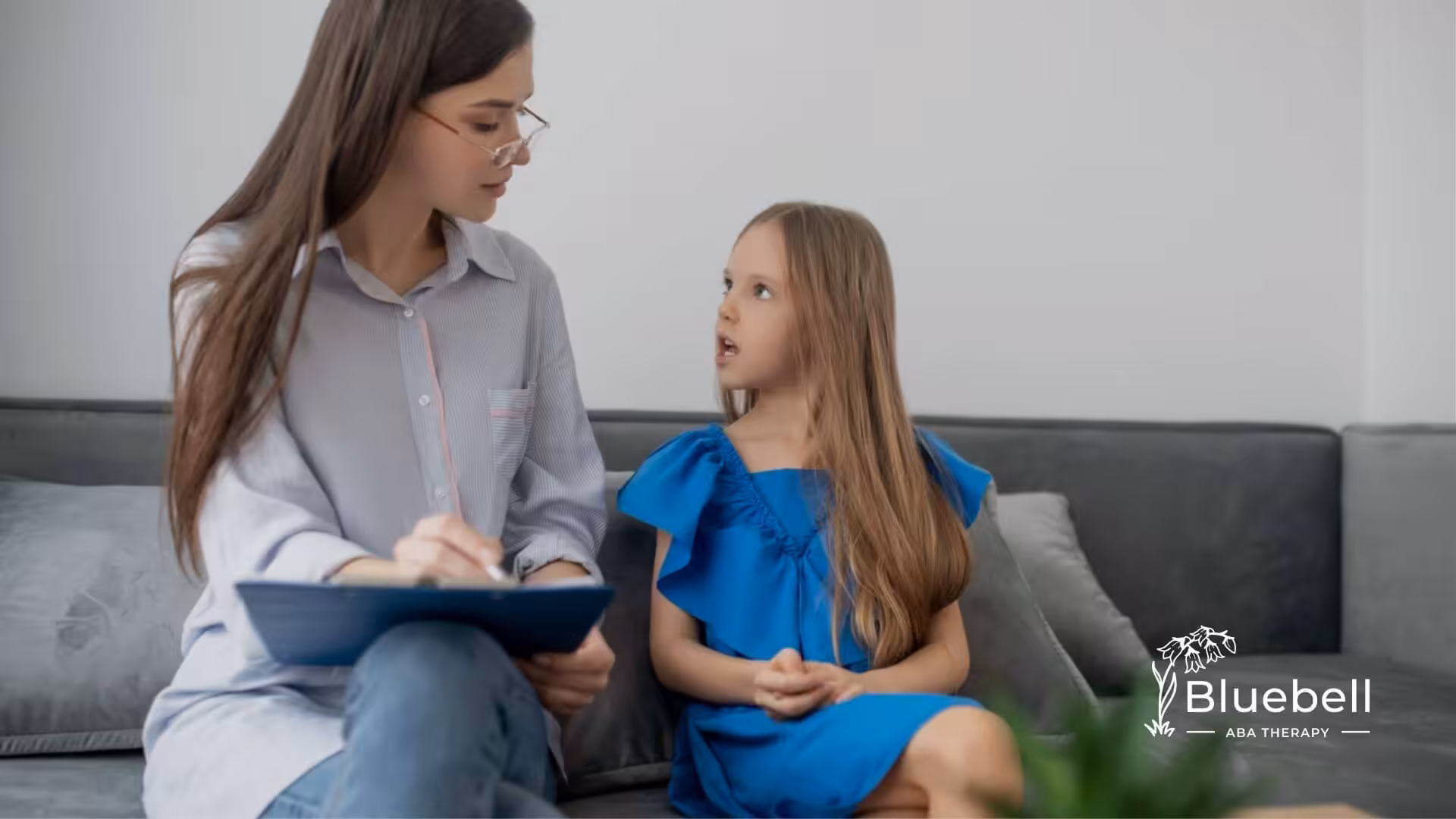 An ABA therapist listens attentively to a young girl with autism sitting beside her on a sofa during ABA therapy.