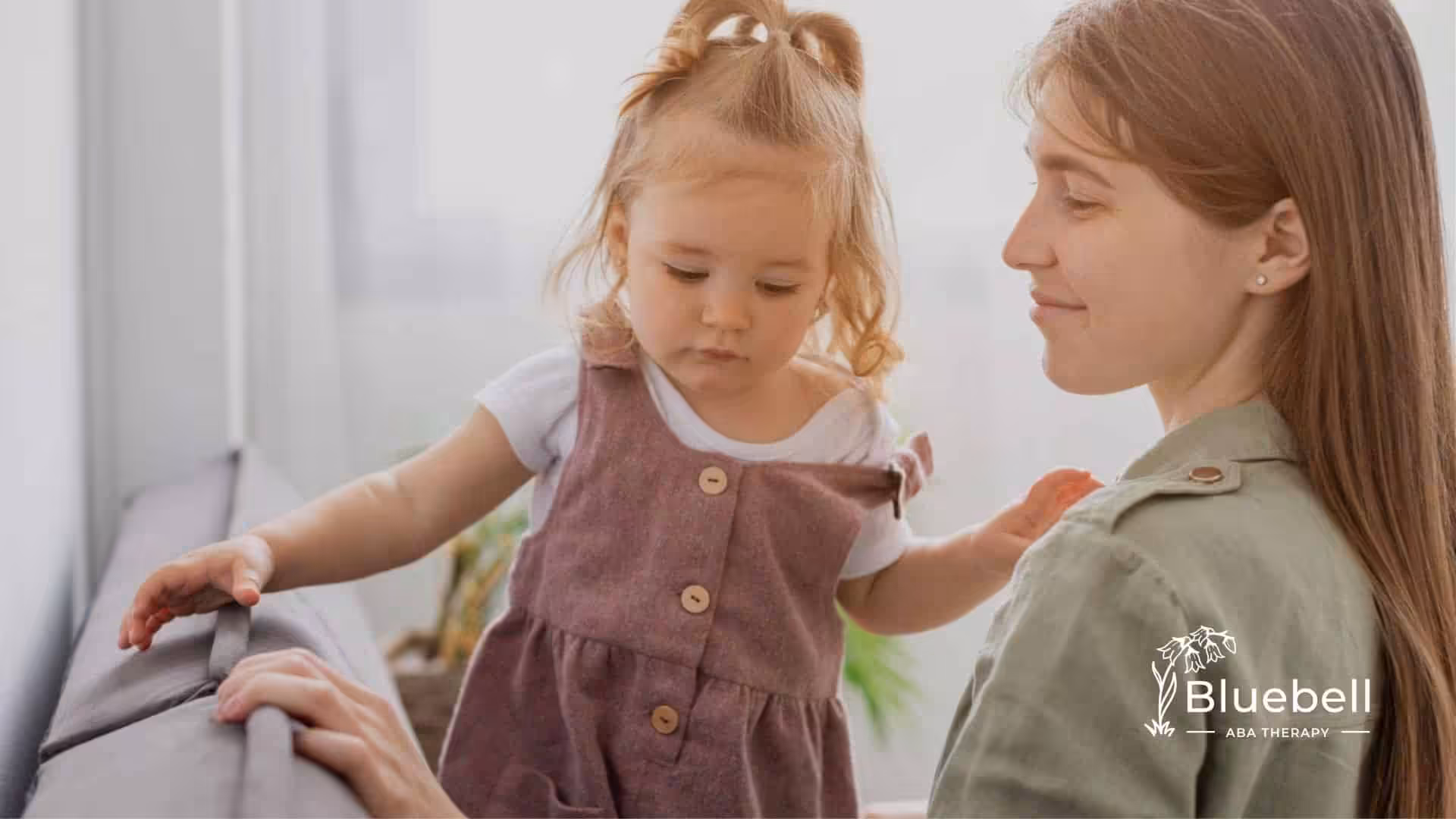 A BCBA supports a toddler with autism standing on a couch, both looking down gently.