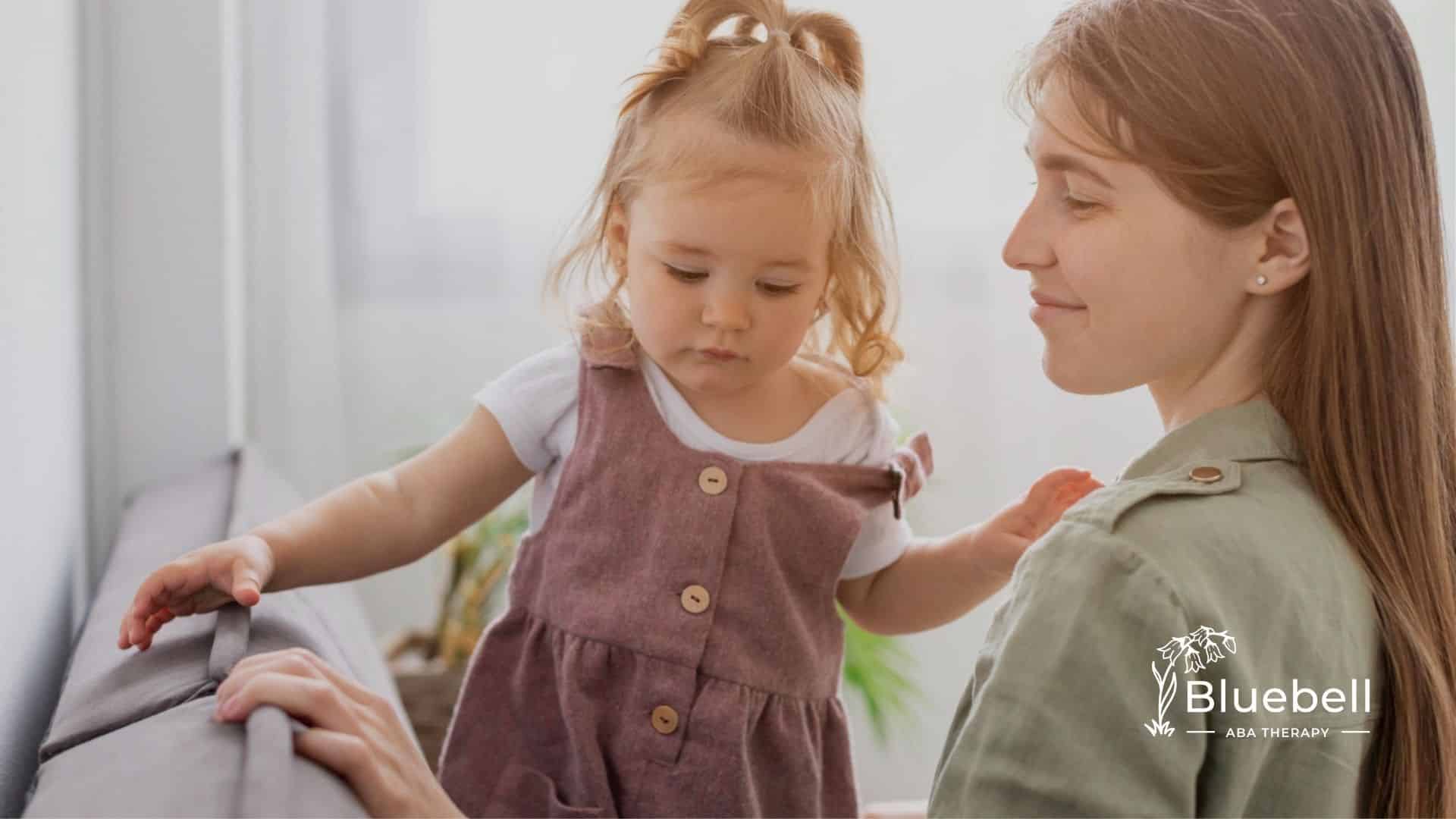 A BCBA supports a toddler with autism standing on a couch, both looking down gently.