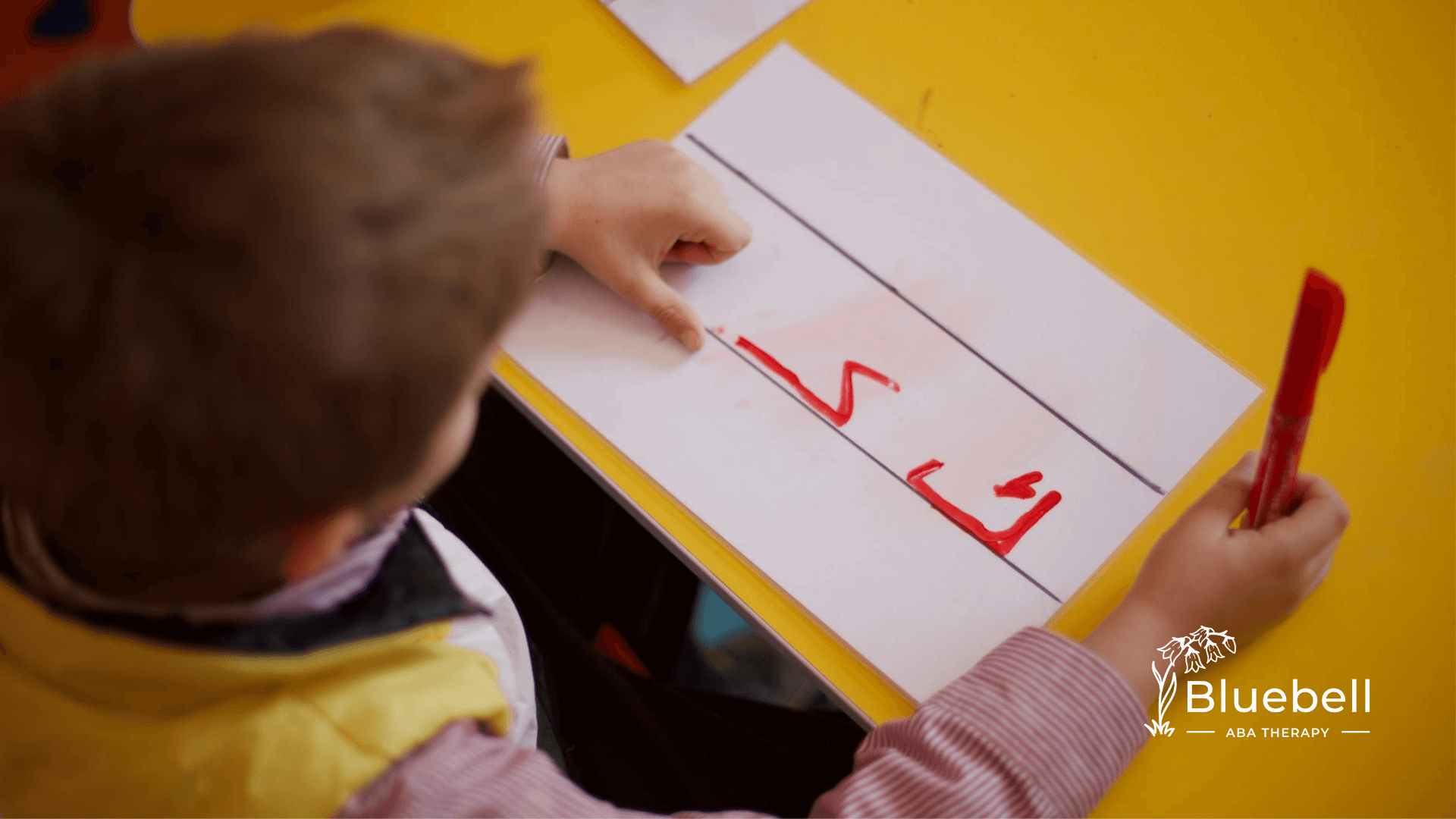 The autistic child in striped shirt concentrates while writing with red marker on lined paper at a yellow table