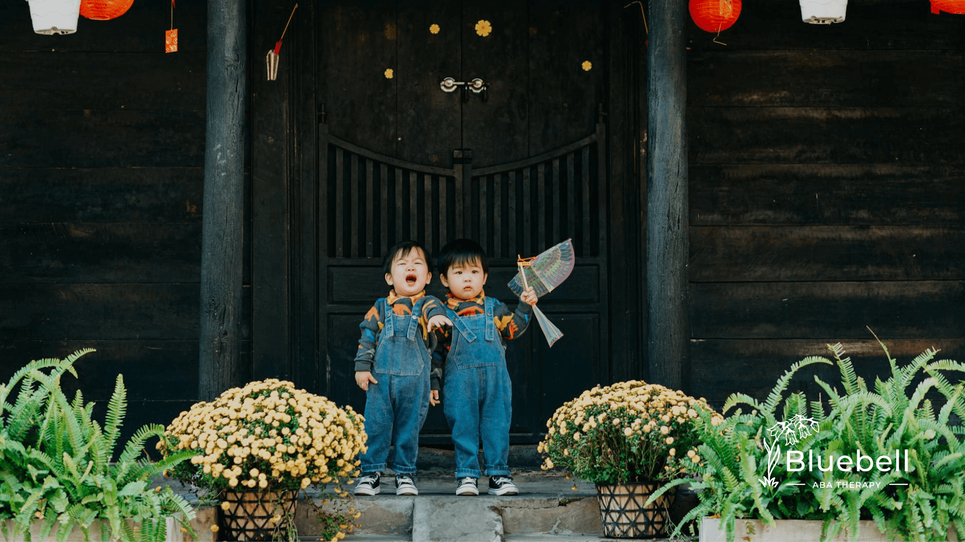Two children in denim overalls pose by a festive wooden building with red lanterns and yellow blooms.