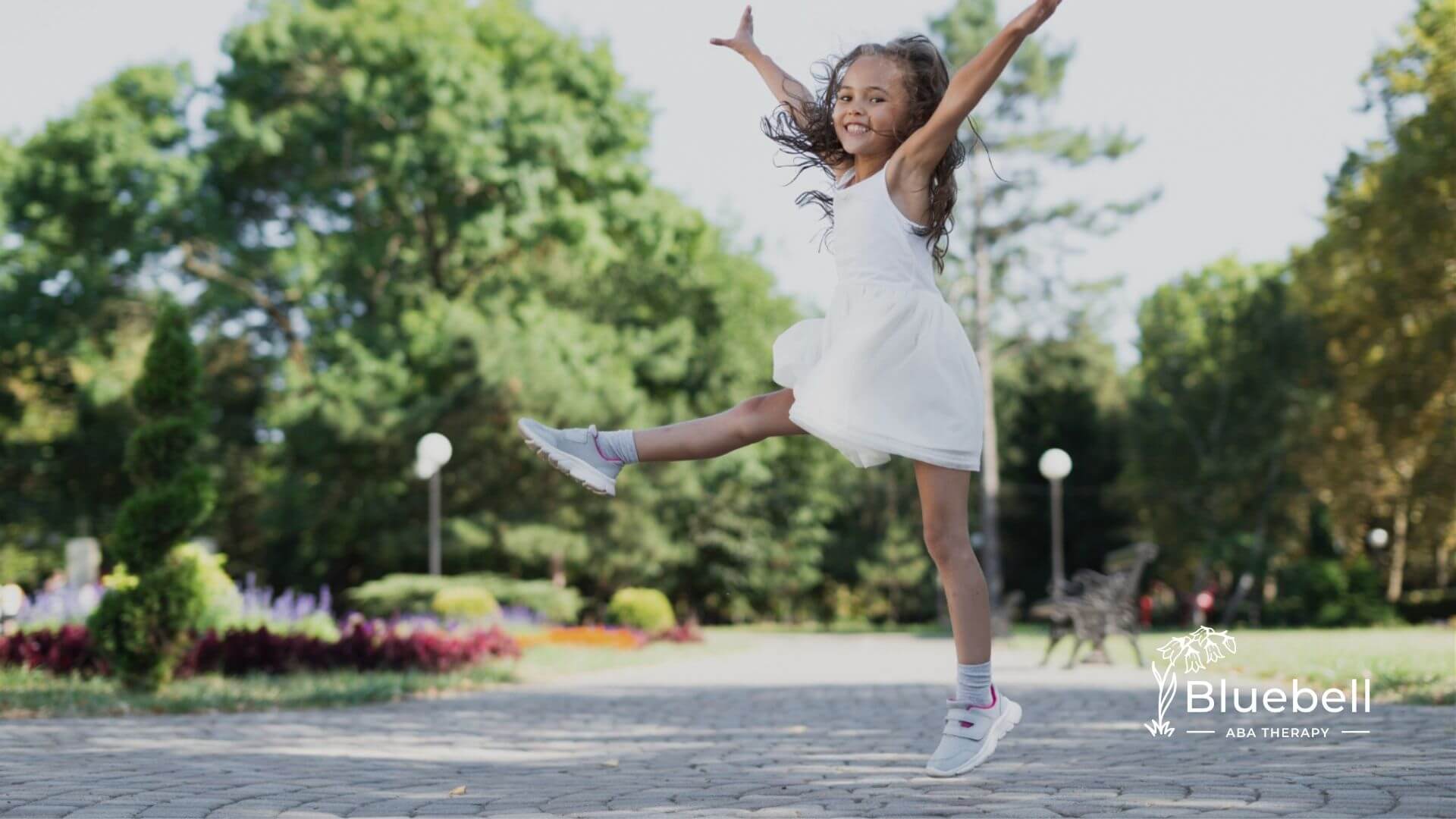 A young girl with autism joyfully jumps in a park, wearing a white dress and sneakers.