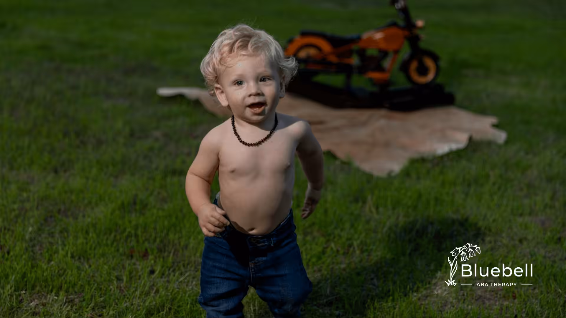 Blonde child in blue jeans walks across grassy field toward a small wooden motorcycle on a decorative rug.