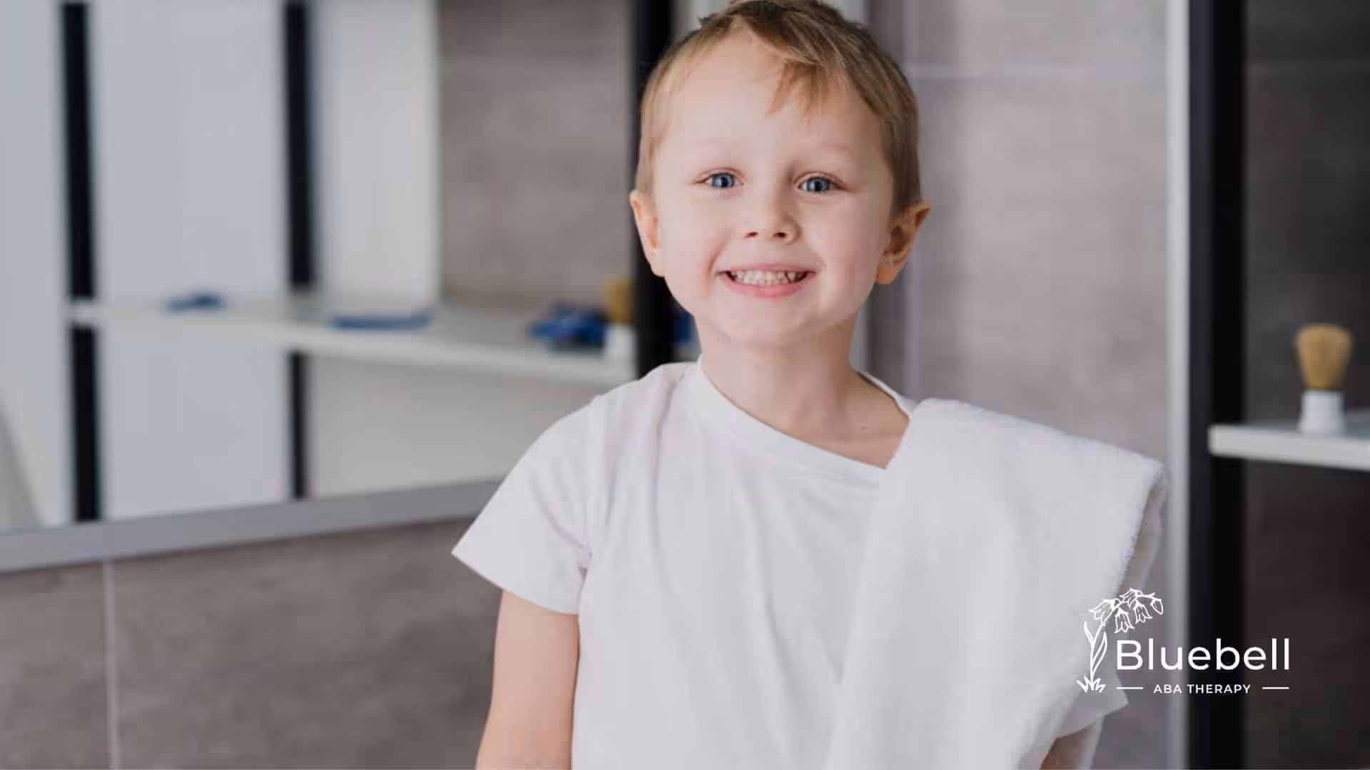 A smiling young boy with autism stands in a bathroom holding a white towel over his shoulder.