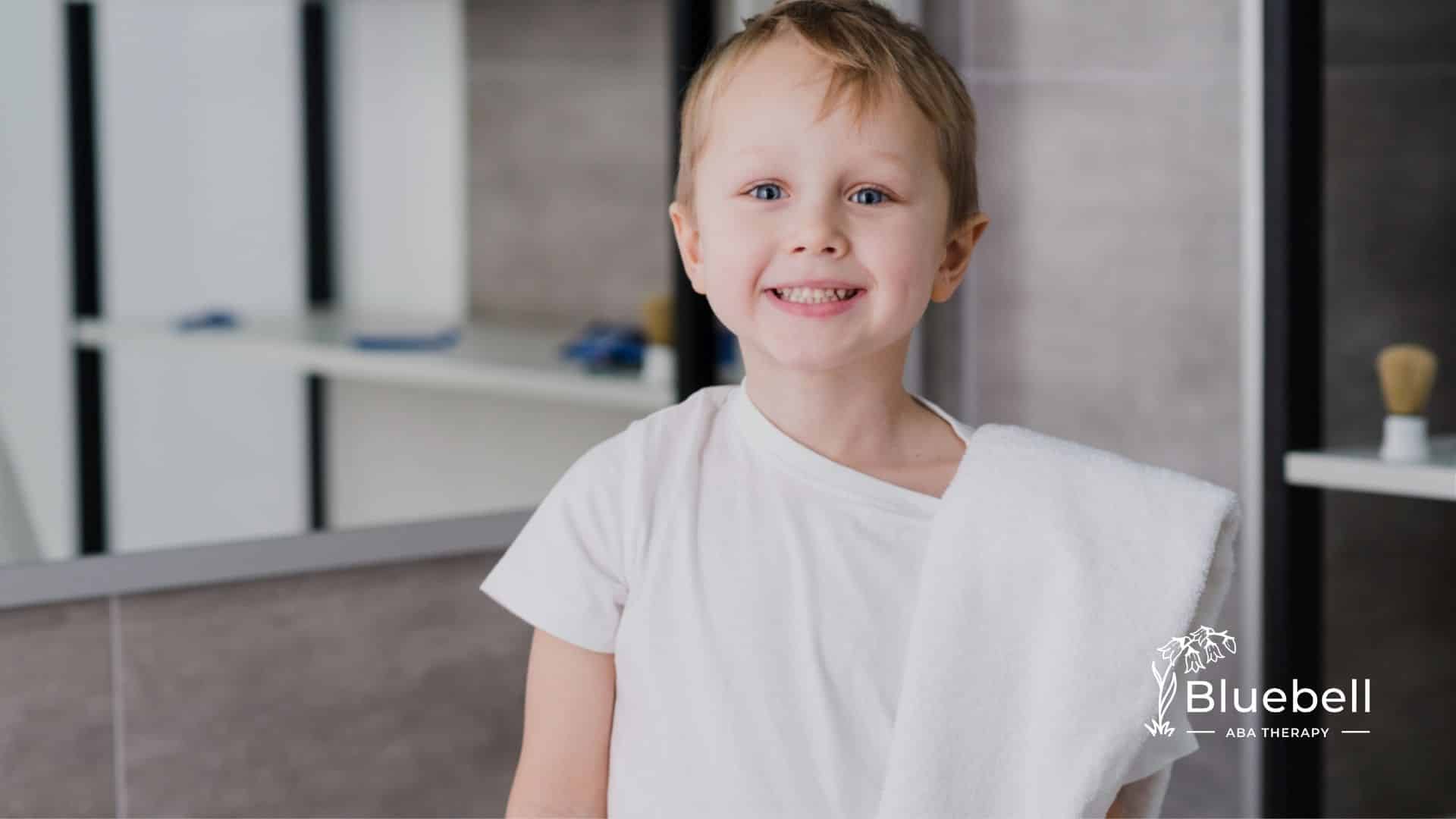 A smiling young boy with autism stands in a bathroom holding a white towel over his shoulder.