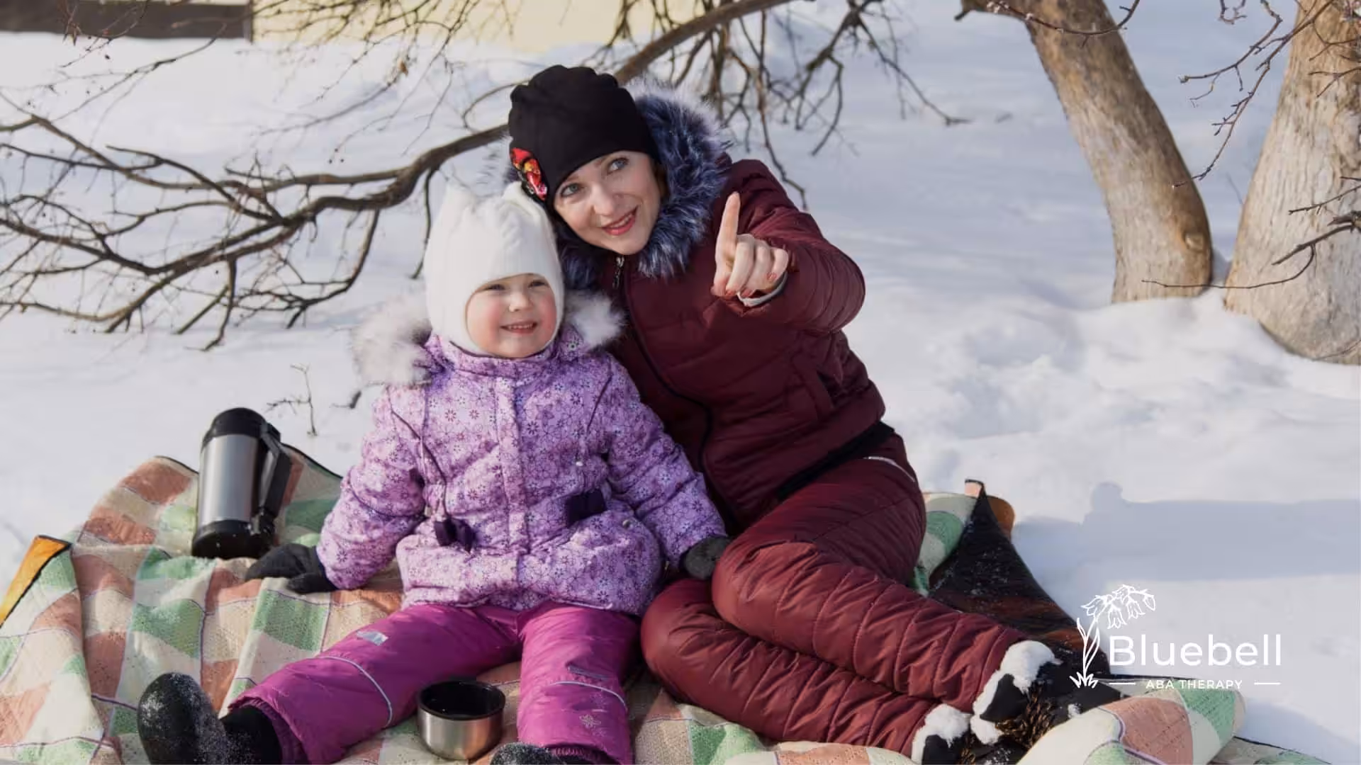 A BCBA and a child with autism sit on a blanket in the snow, with the BCBA pointing at something.