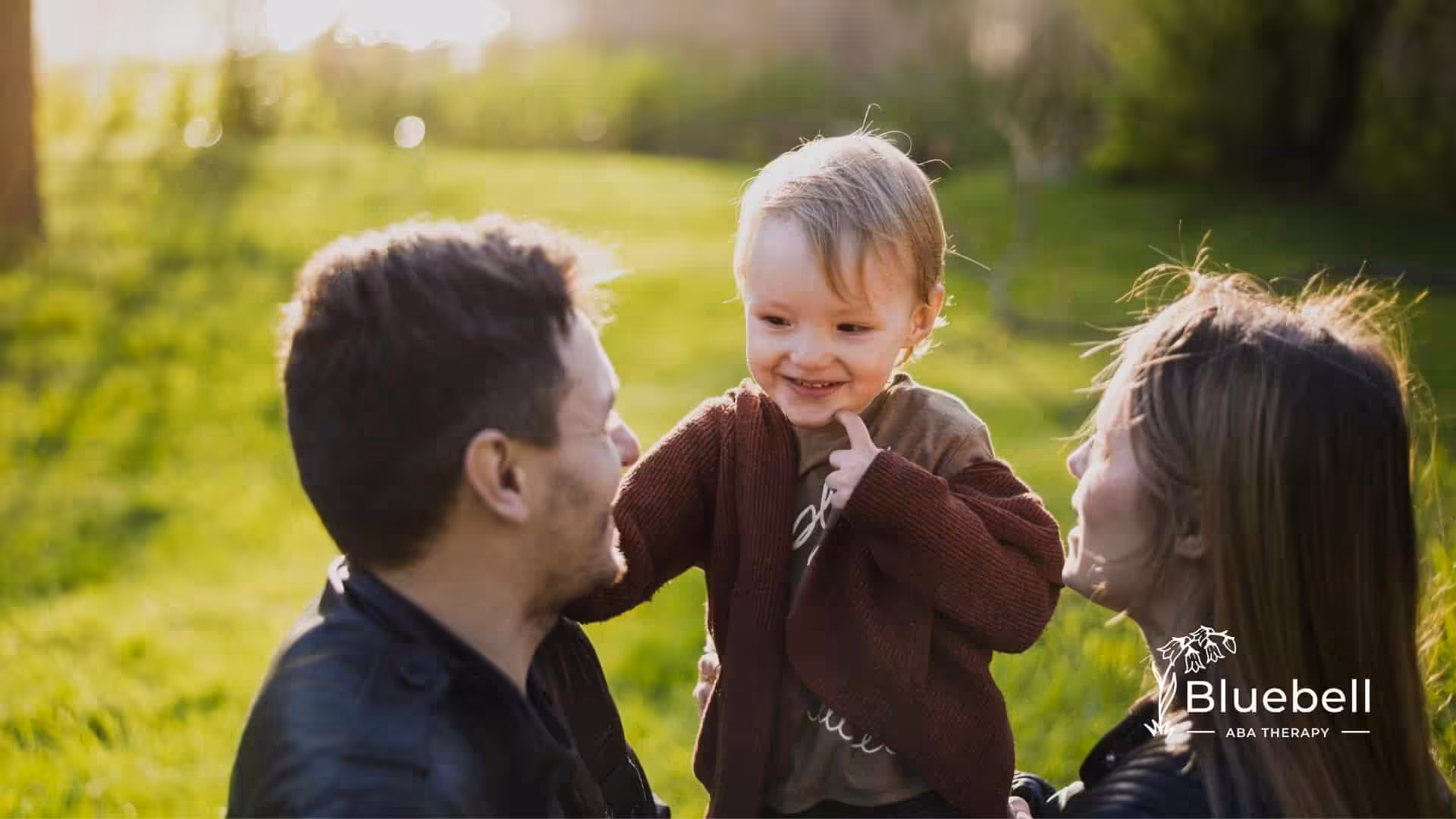 Smiling autistic toddler interacting with both parents outdoors, showing strong attachment, connection, and family support.