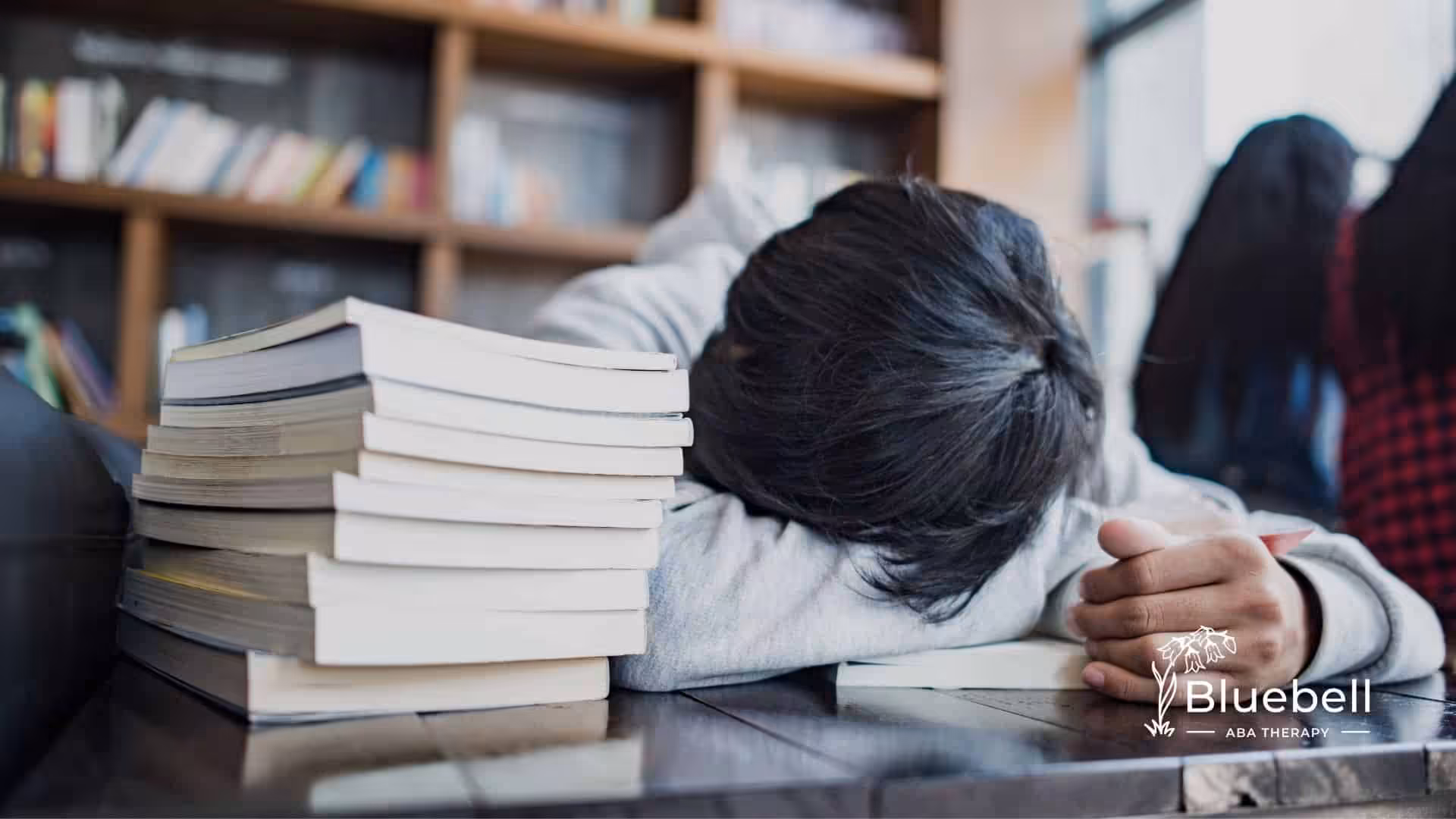 Exhausted ABA student resting head on desk beside stacked textbooks, showing burnout and stress from intensive exam studying.