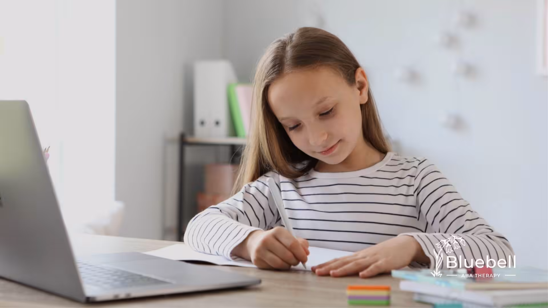 Young child with autism writing in a notebook at a desk with a laptop nearby.