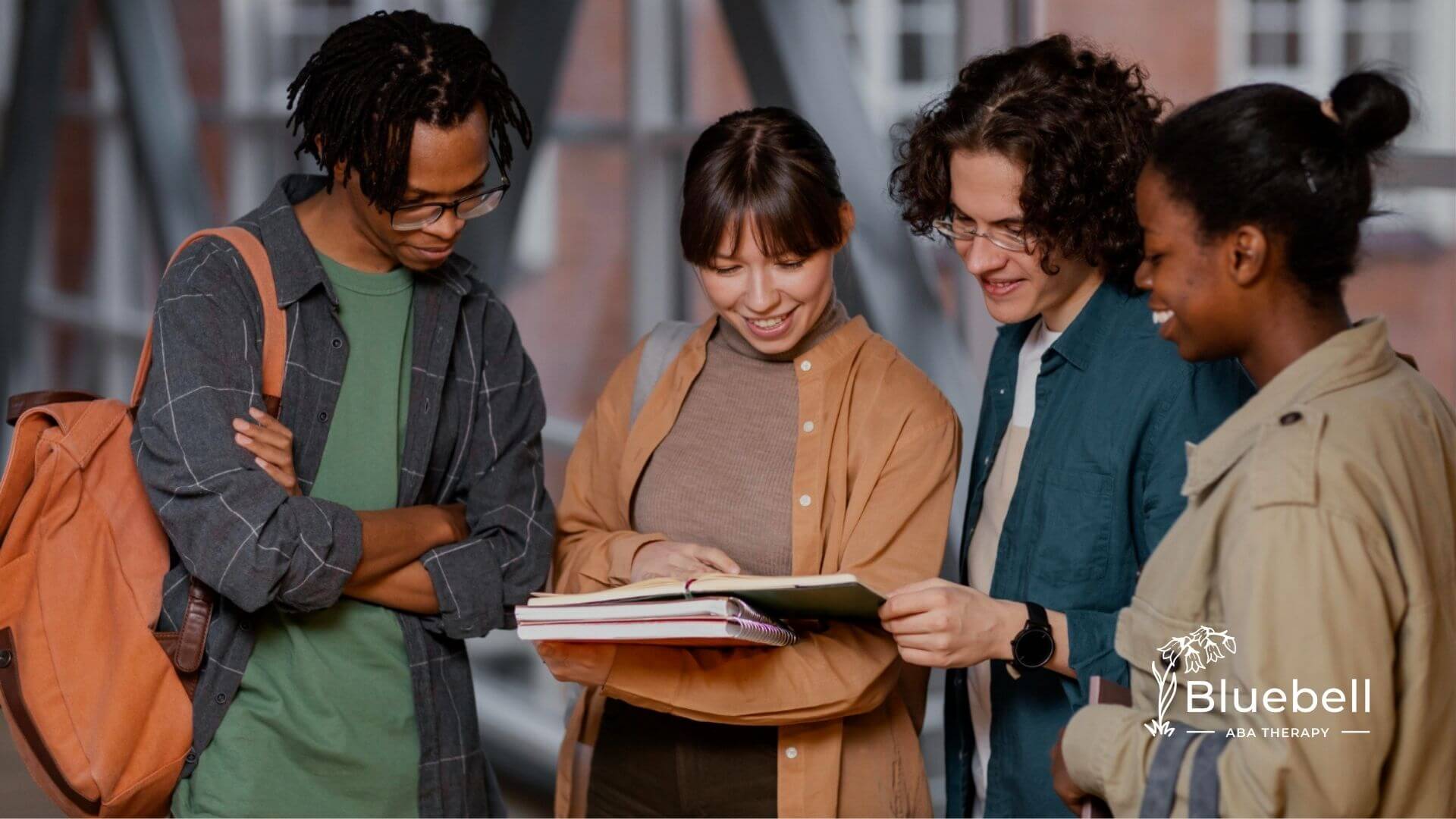 Four college students standing together outdoors, smiling and looking at an open notebook.