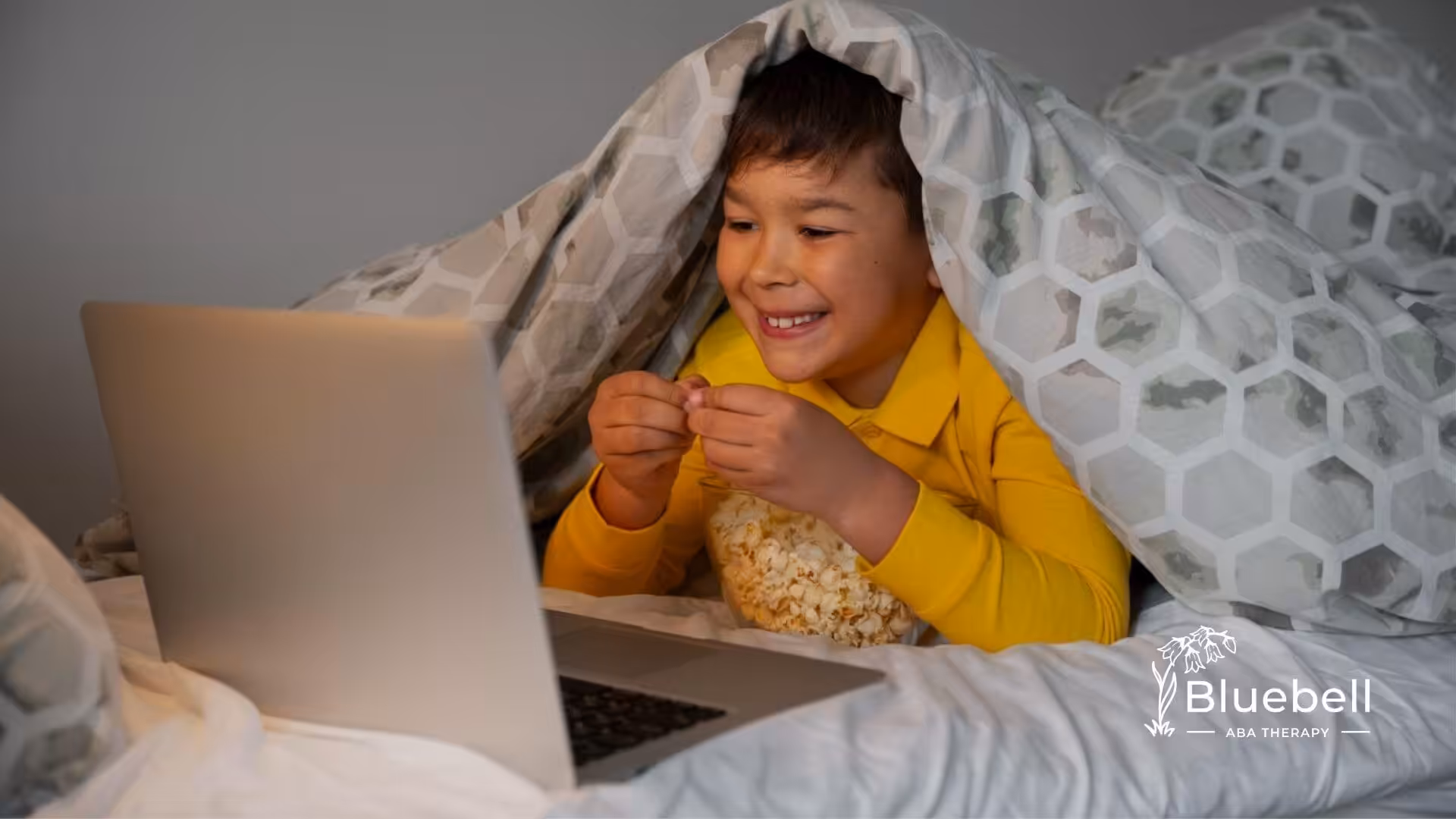 Child with autism watching a laptop under a blanket with popcorn.