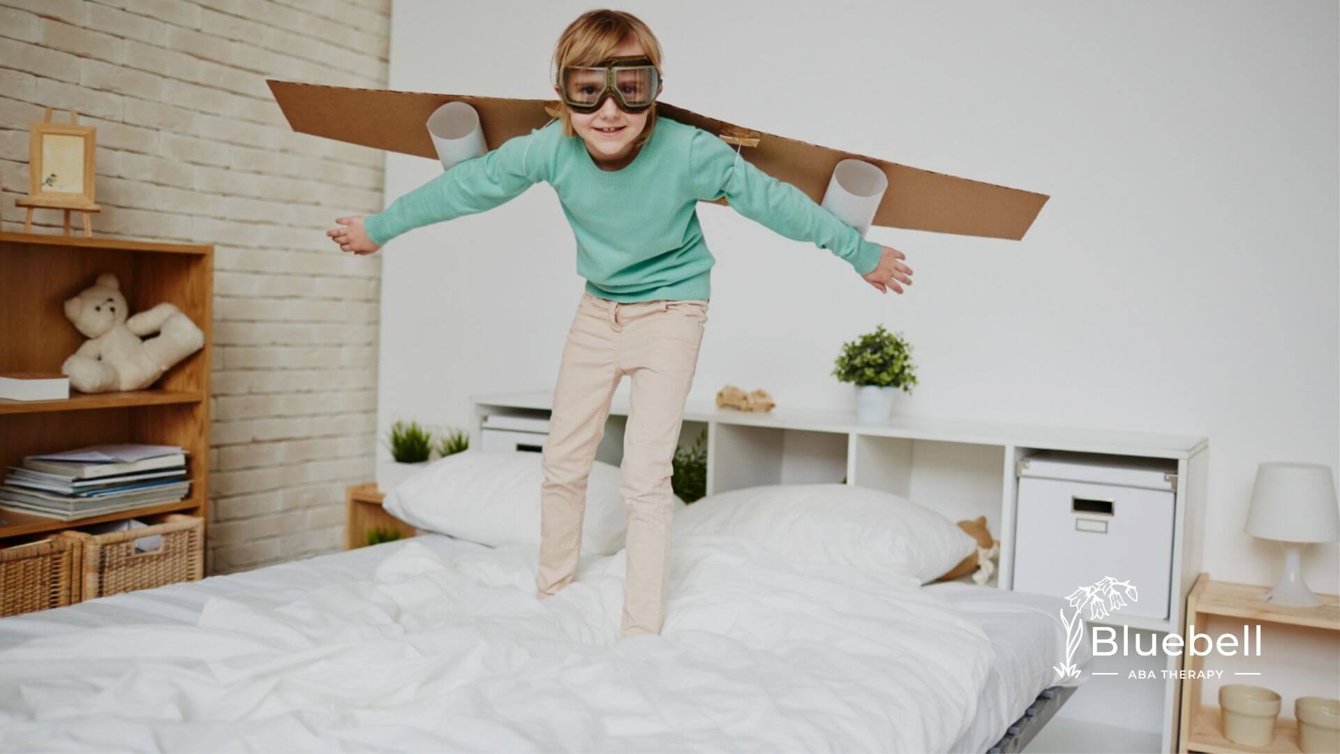 Child wearing goggles and cardboard wings pretending to fly while standing on a bed in a bedroom.