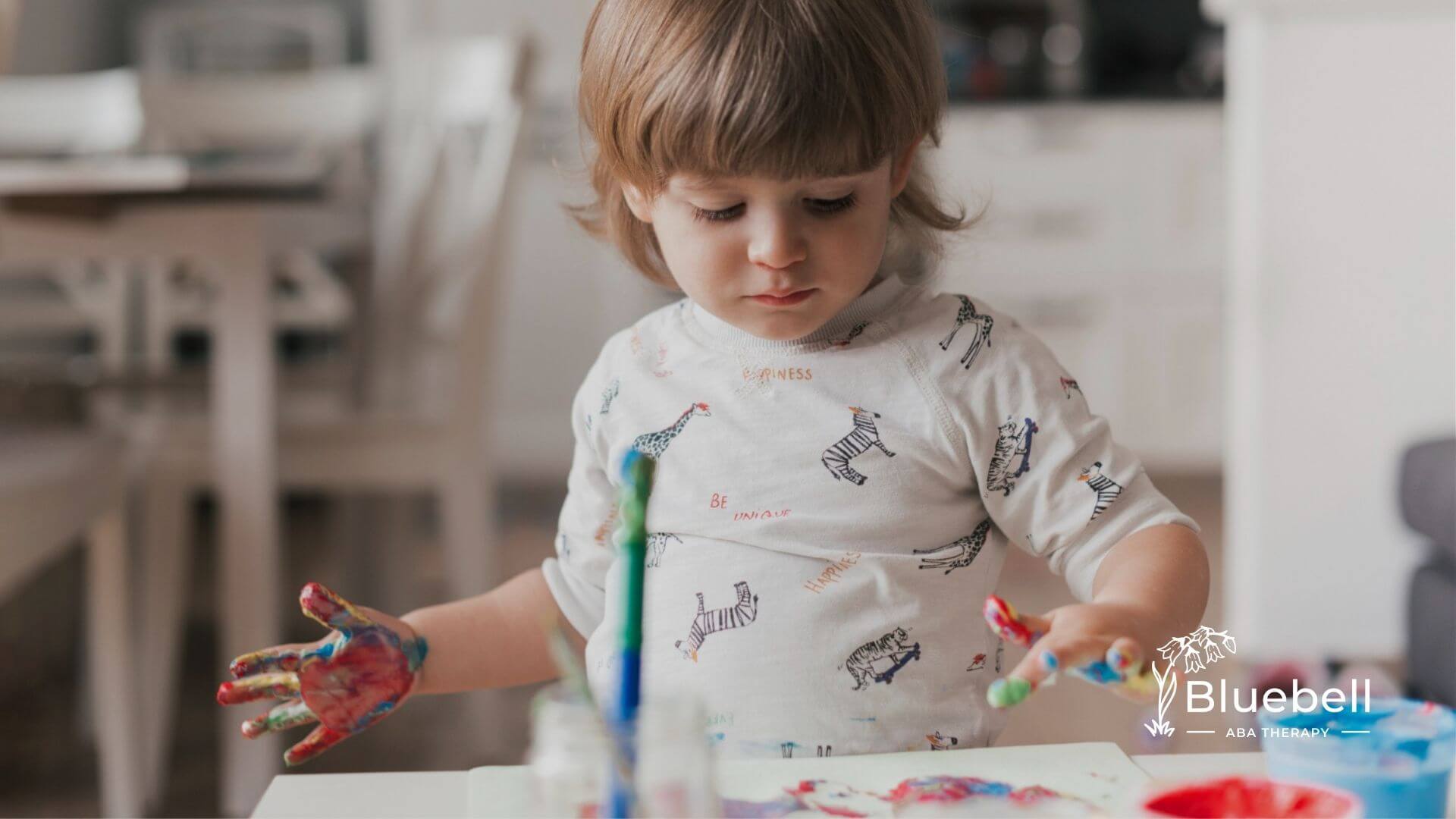 Autistic toddler with paint-covered hands looking down at colorful artwork on a table.
