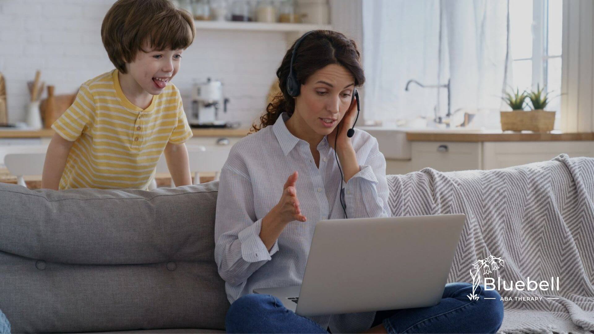 BCBA wearing a headset working on a laptop while a young child stands behind her on a couch.