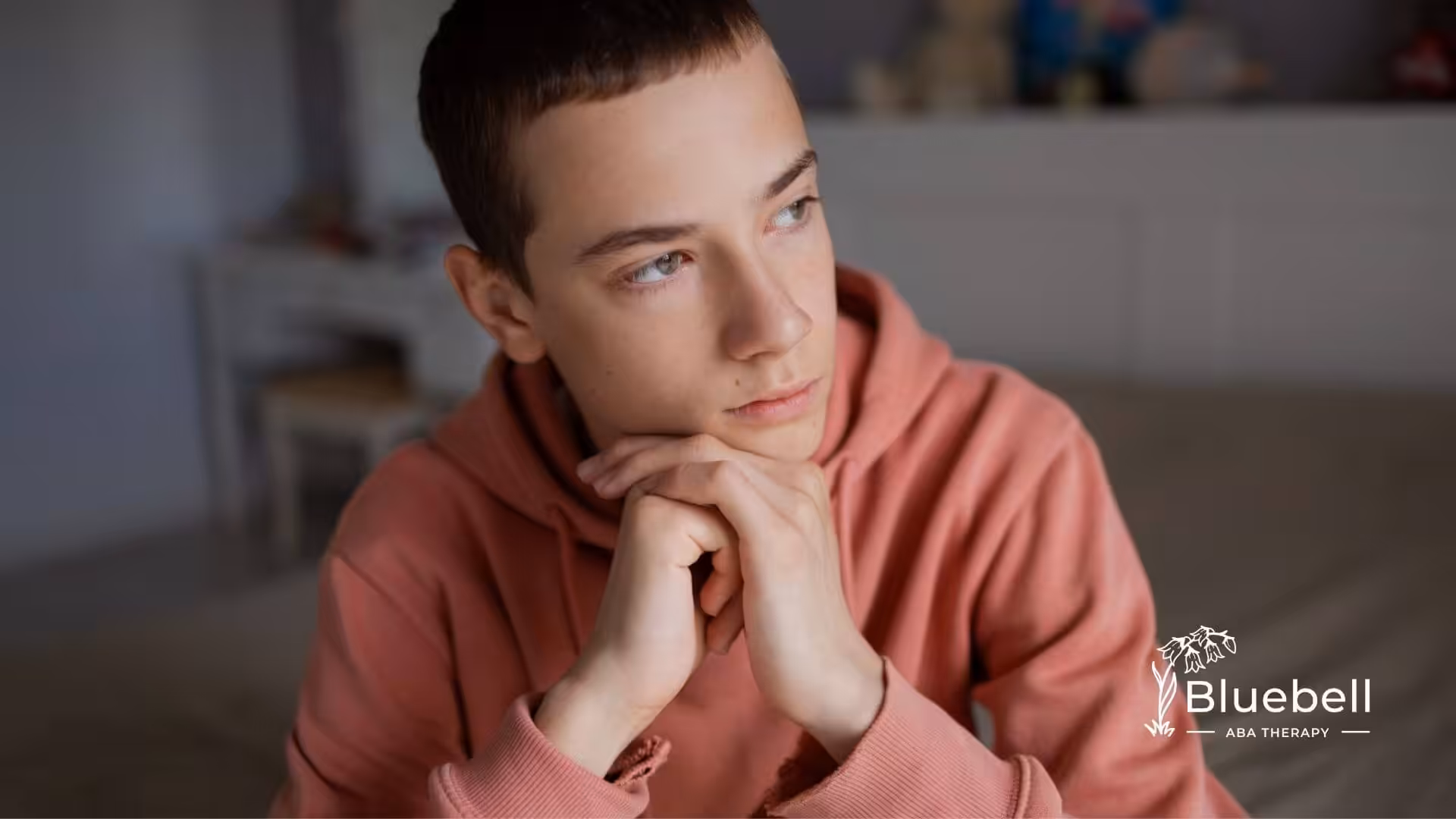 Autistic teenager with a shaved head resting their chin on their hands and looking thoughtfully to the side.