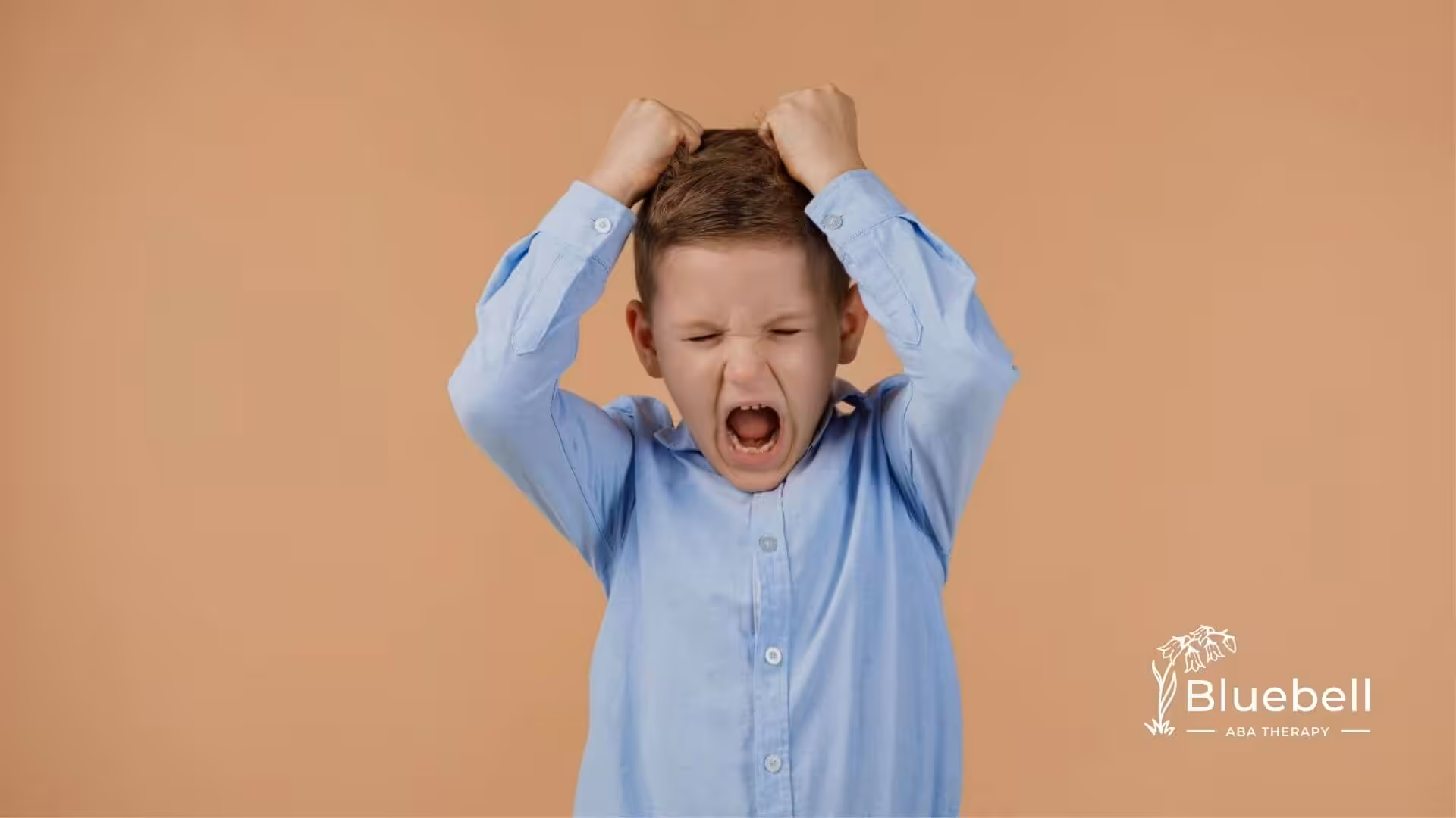Young boy with autism pulling his hair and yelling in frustration against a plain background.